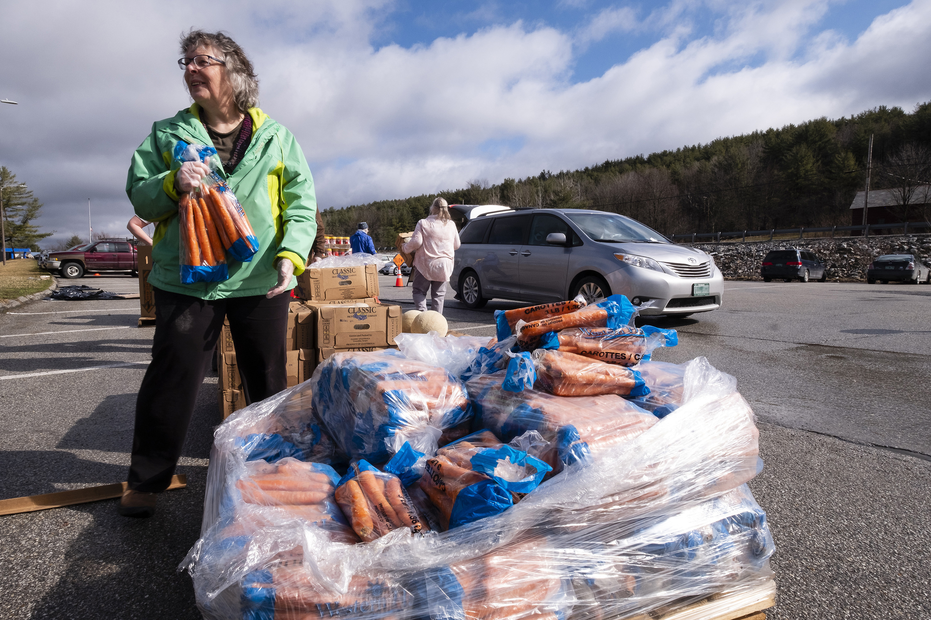 An older white woman is seen holding a large bag of carrots in a rural parking lot. In front of her is a pallet of carrots, bagged in bundles. Behind her, cars are seen lined up. In the background, a dense forest of evergreen trees are seen. People are dressed in light coats.