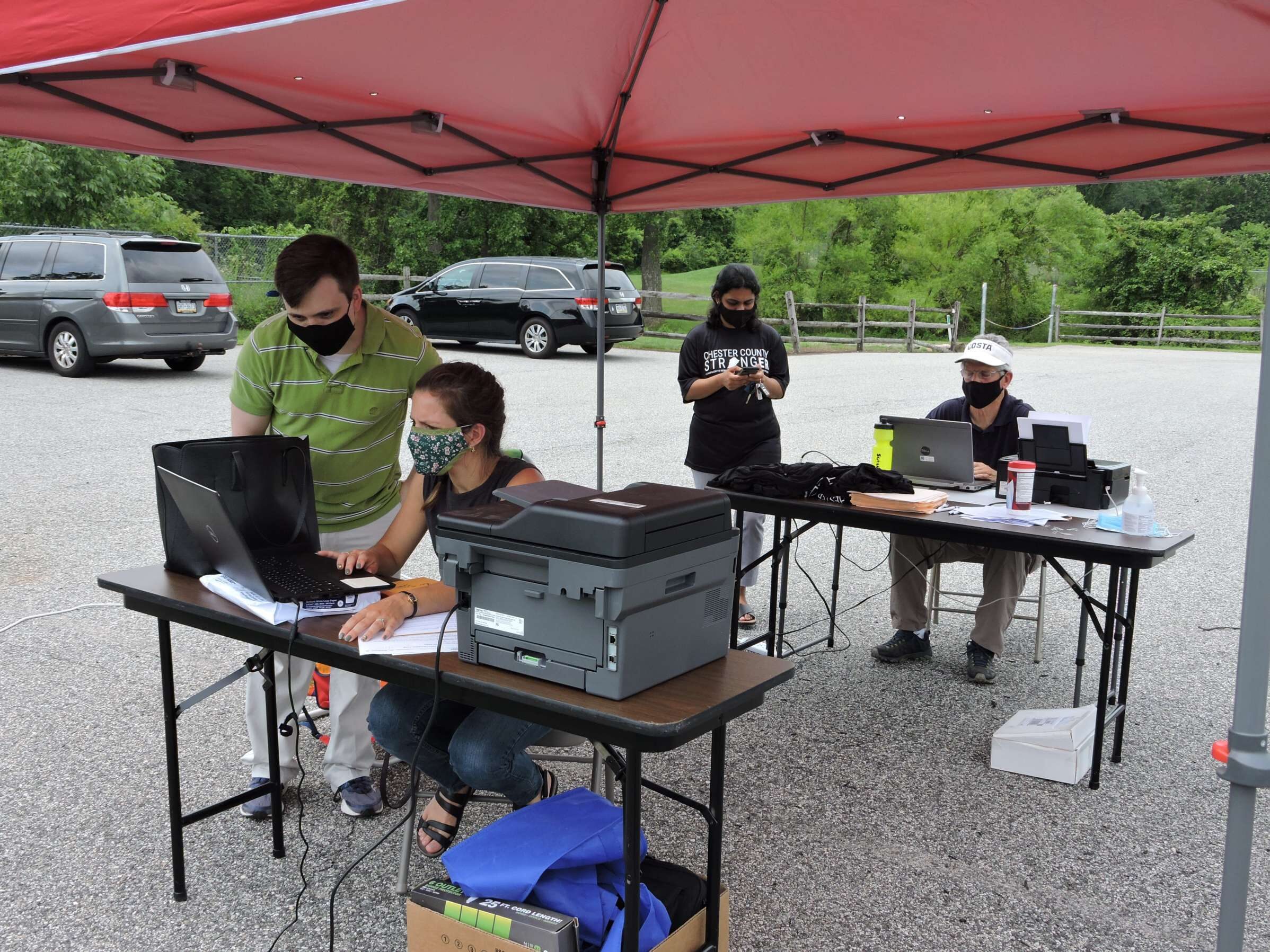 Four people are seen outside in a parking lot under a red tent pop-up tent. In the background, the trees are green. Two of the people,  one man, one woman, sit at two different tables, each with a laptop and a printer visible. The other two stand next to the people at the table, one at each. They appear to be involved in conversations.