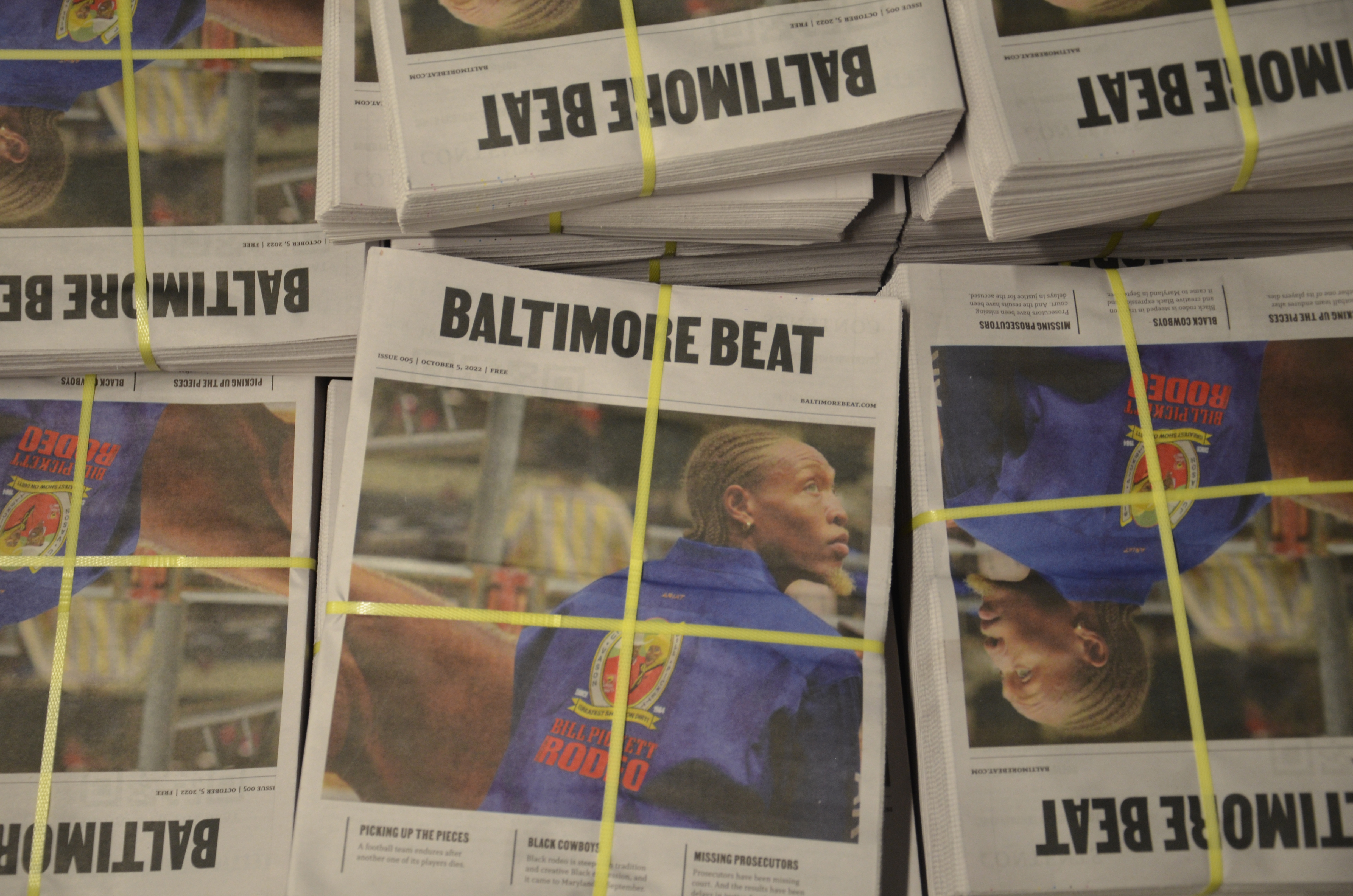 Stacks of the October 5, 2022, edition of the Baltimore Beat newspaper are seen from above, still bundled with strips of plastic. The cover image features a brown-skinned man with light-colored dreadlocks and goatee photographed from behind. The man is looking into the distance off camera right. He is wearing blue jacket bearing the words and logo of “Bill Pickett Rodeo.”