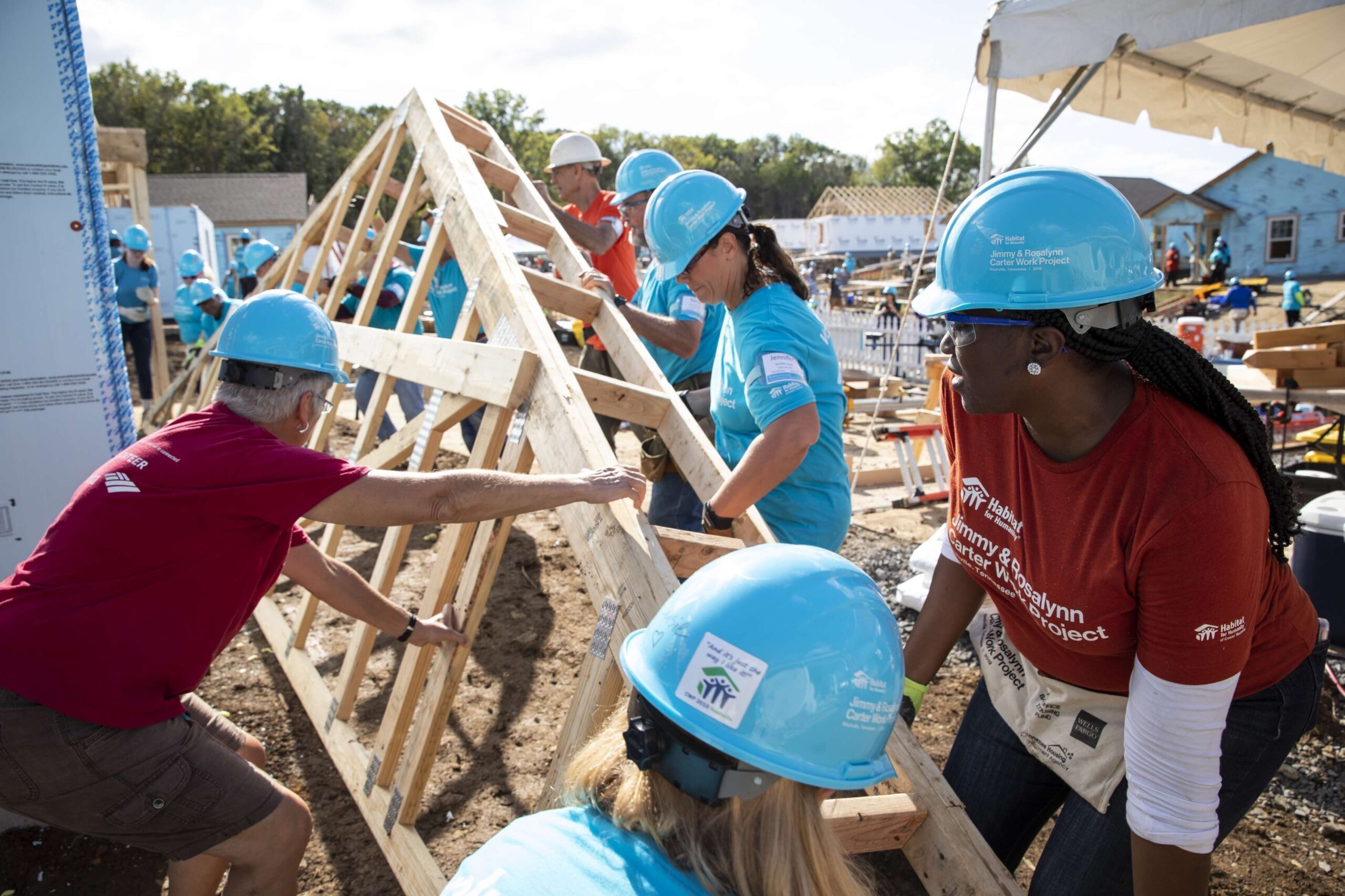 Roughly a dozen people are seen lifting a wooden roof truss on a construction site. The people are all wearing matching blue construction helmets.