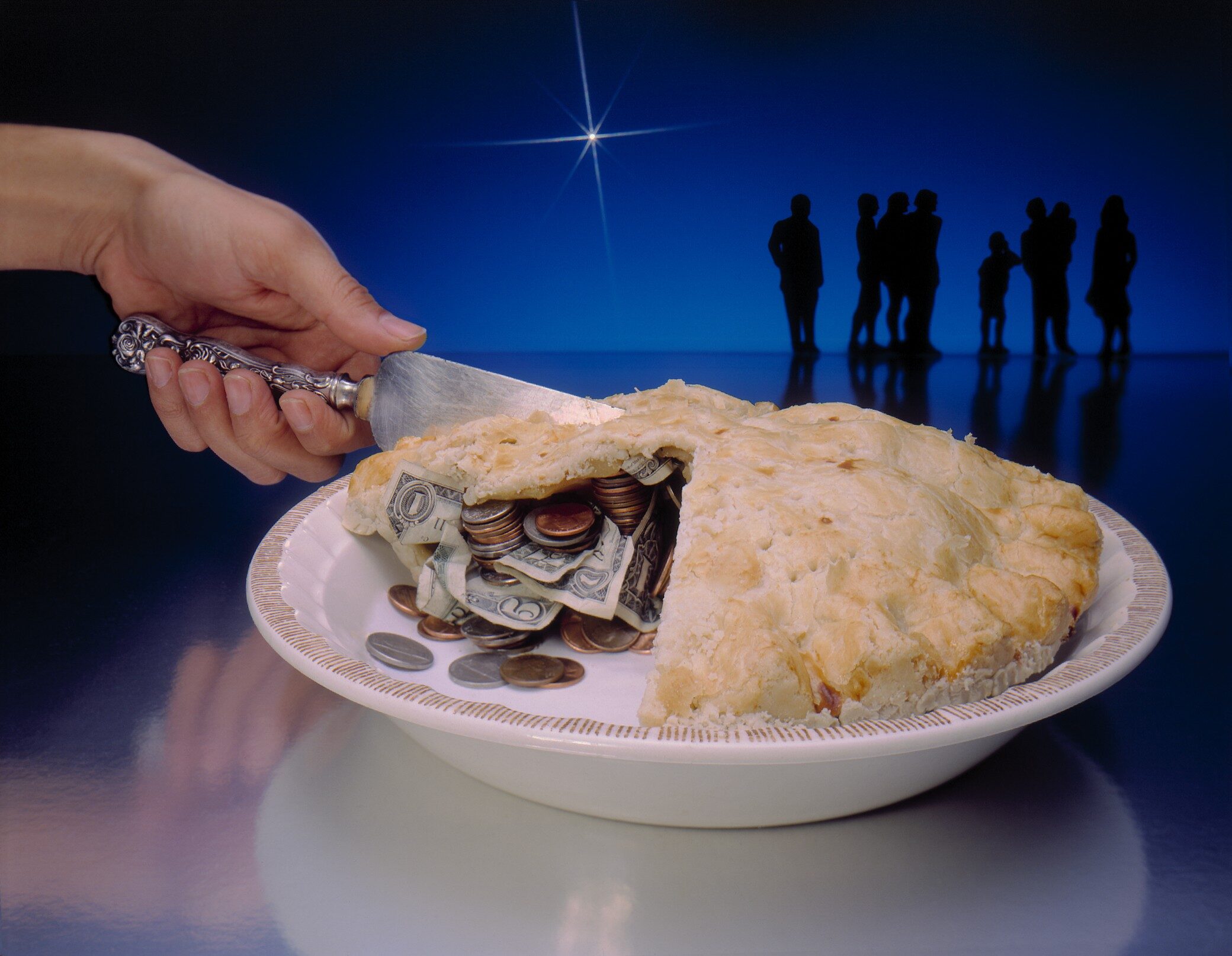 Hand cutting into a baked pie filled with money. There  are silhouettes of a group of people standing in the background. The background is blue with a  bright cross star.