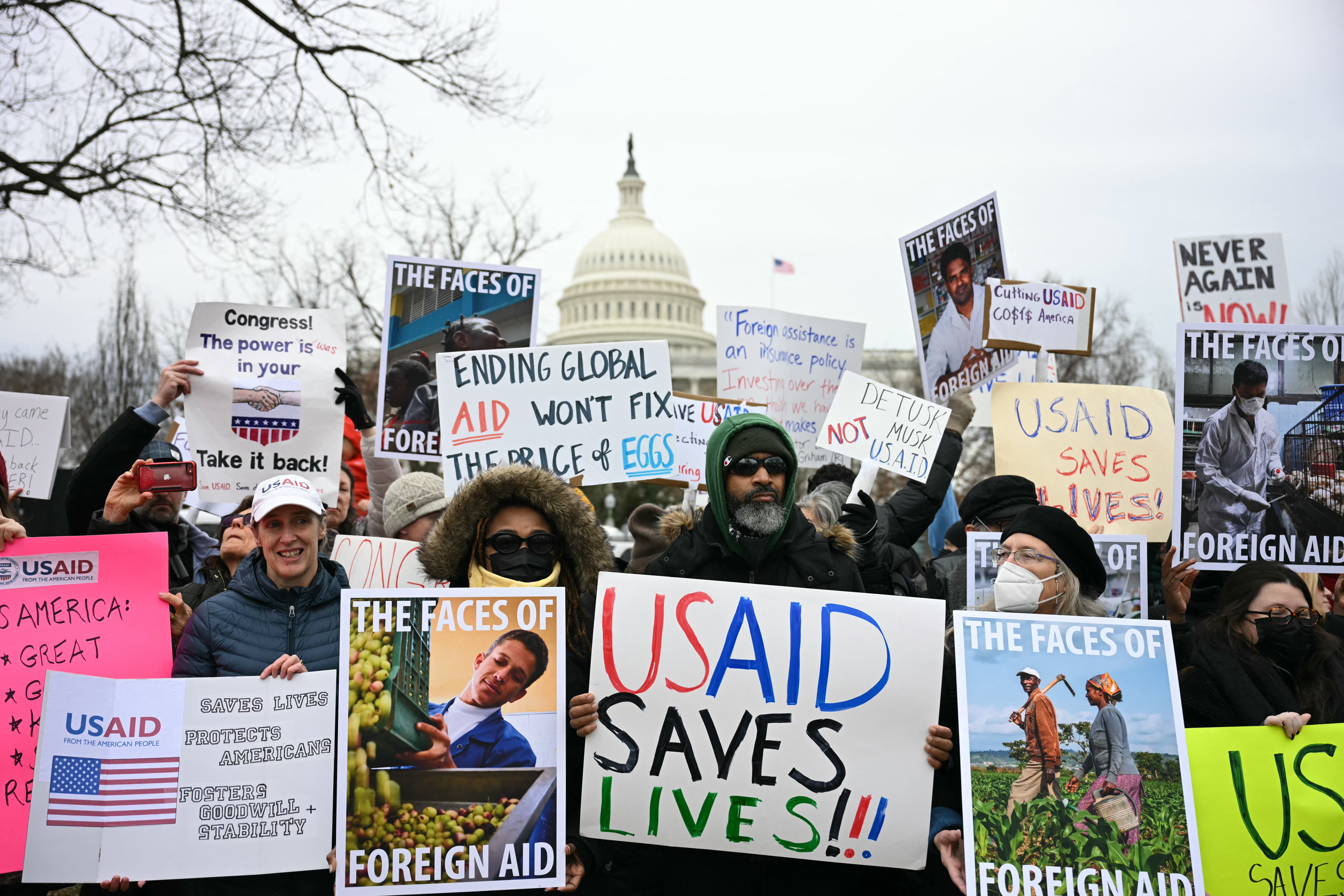 A group of of protestors hold signs reflecting their anger about Elon Musk's plan to shut down USAID. Sign messages include "USAID saves lives" and "the faces of foreign aid".  