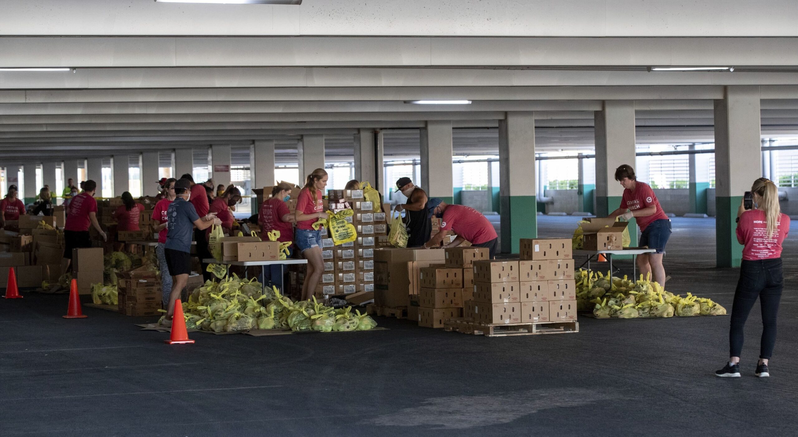 A group of dozens are seen in a parking garage working around pallets of boxes and piles of yellow bagged goods. 