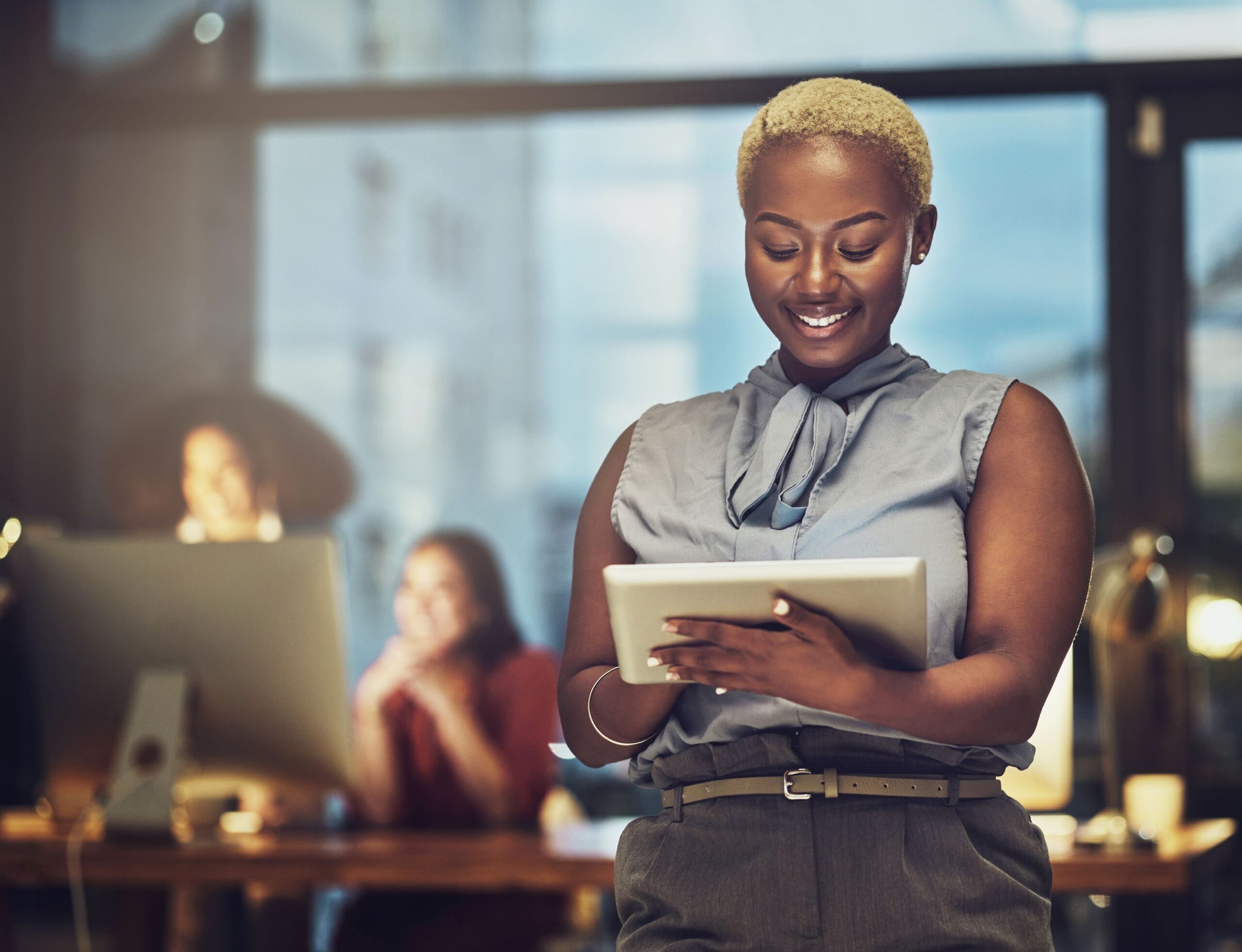 A Black woman in stylish blouse stands in the foreground smiling and looking down at a tablet while coworkers happily work in the background around a large Apple monitor. 