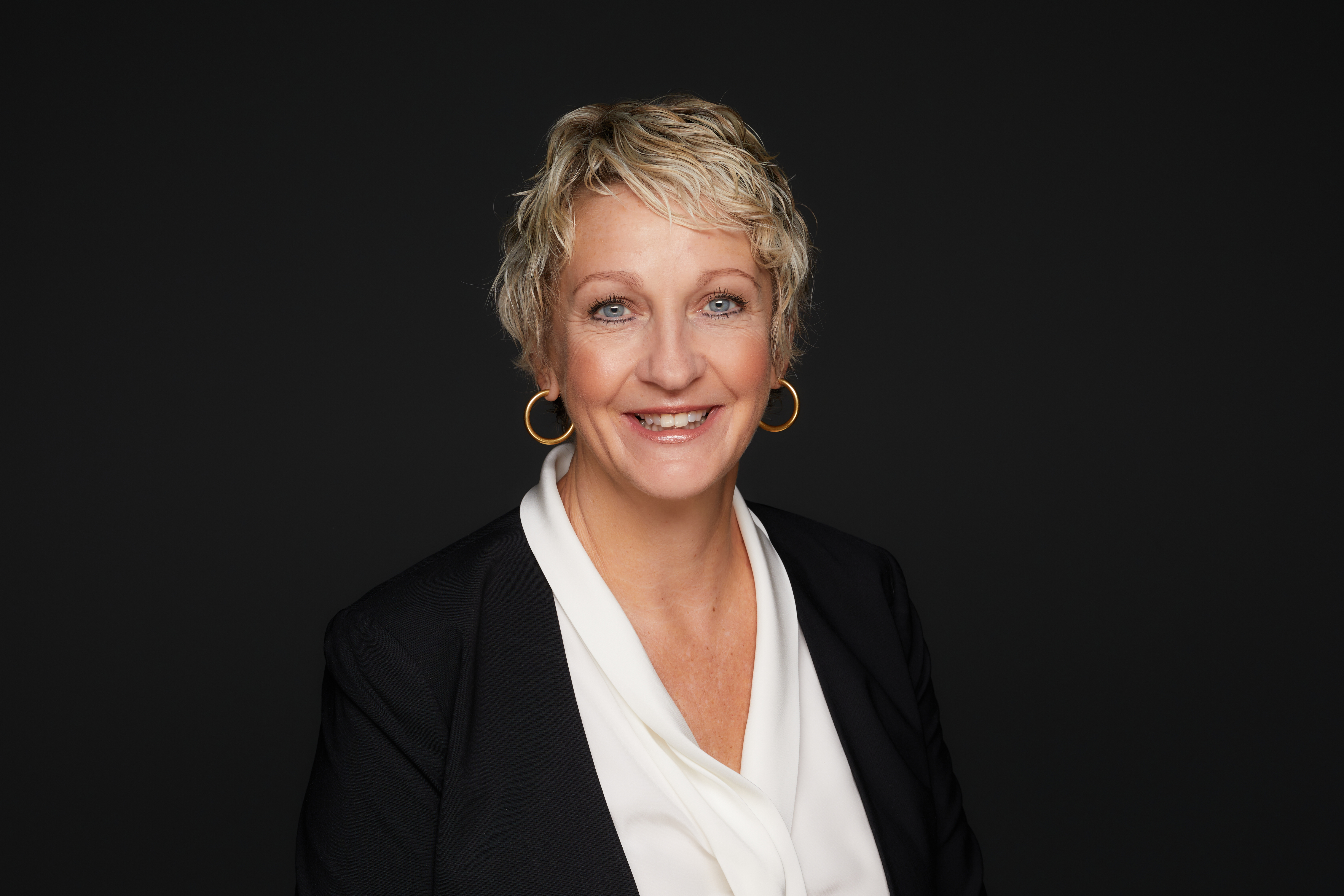A portrait shows a smiling white woman with short blonde-highlighted hair dressed in professional attire. She wears a set of large, round gold earings, a white blouse and a dark jacket. 