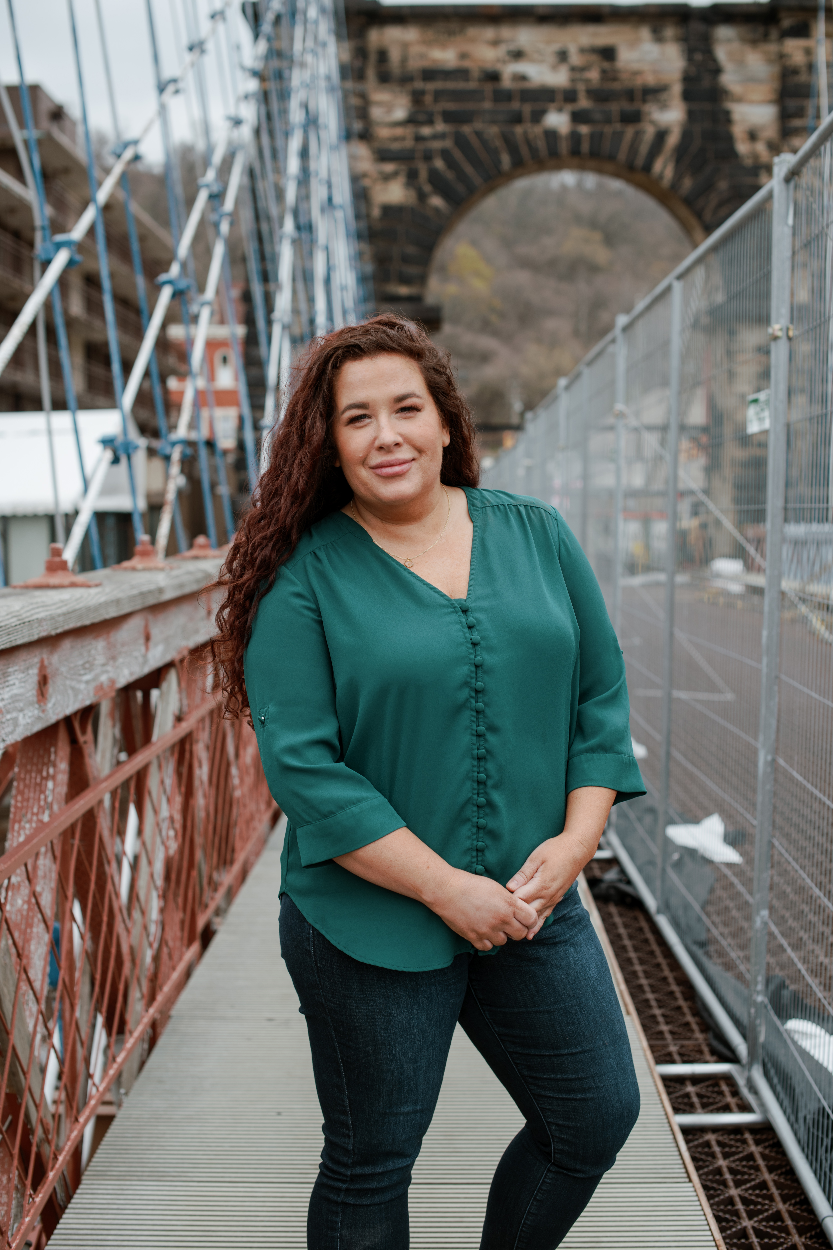 Dressed in a green blouse and blue jeans, a white woman with long brown curly hair poses for a portrait on the sidewalk of an old suspension bridge, with a masonry arch and supporting cables visible in the background. 