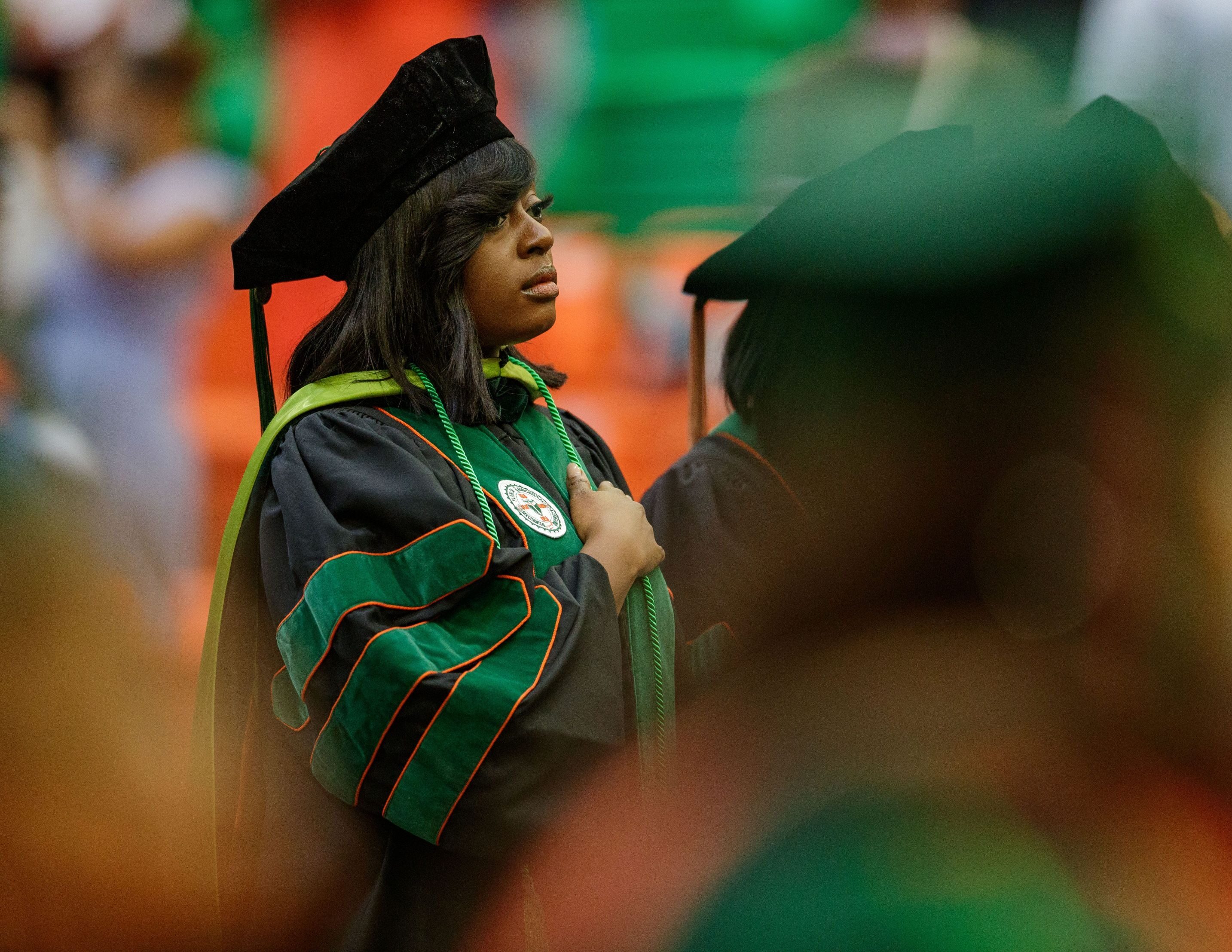 A black woman, dressed in graduation regalia, stands with her hand over her heart during a graduation ceremony.