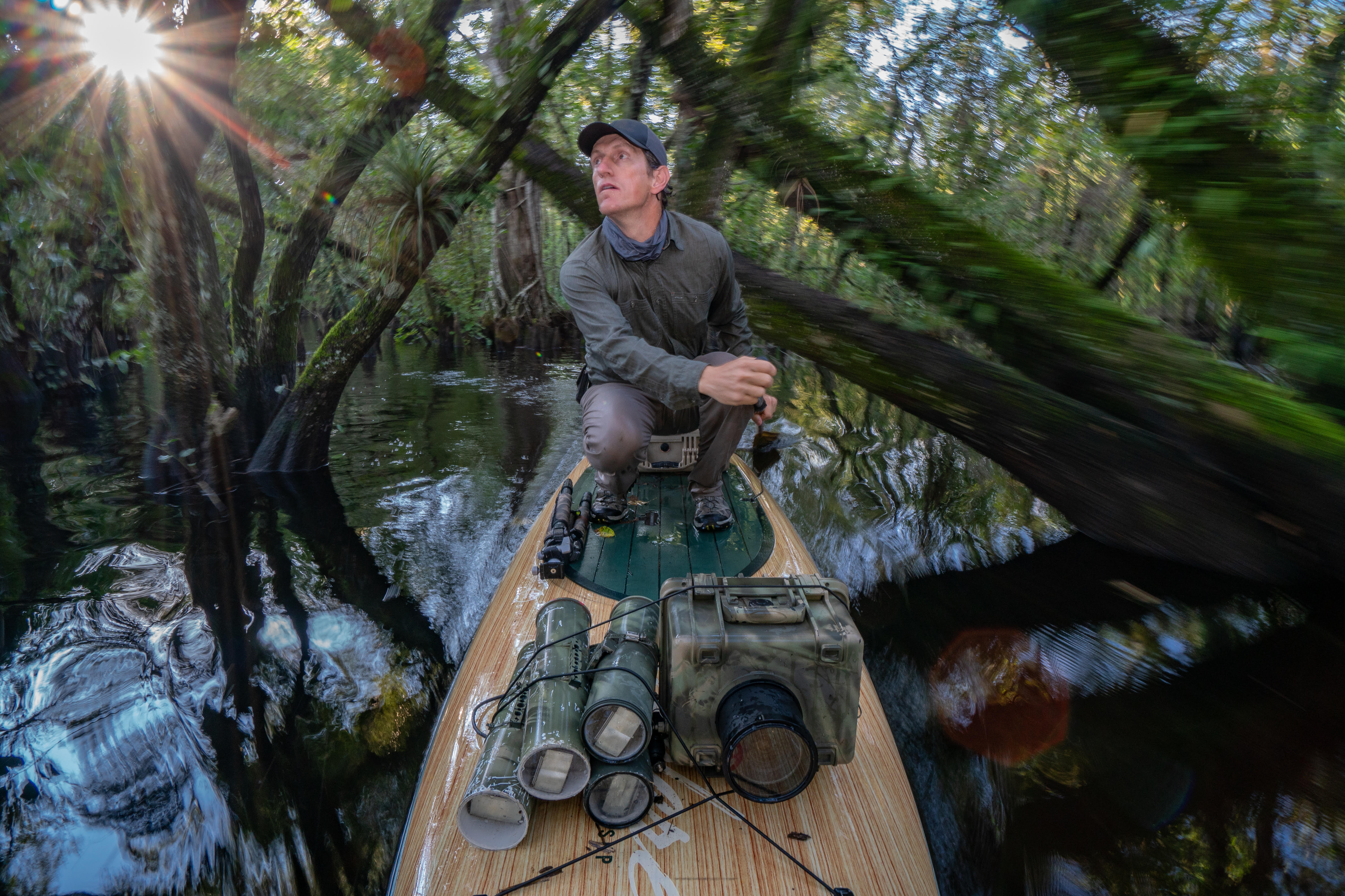 A white adult man guides a paddleboard through a swamp. His boat is laden with camouflaged photographic equipment. 