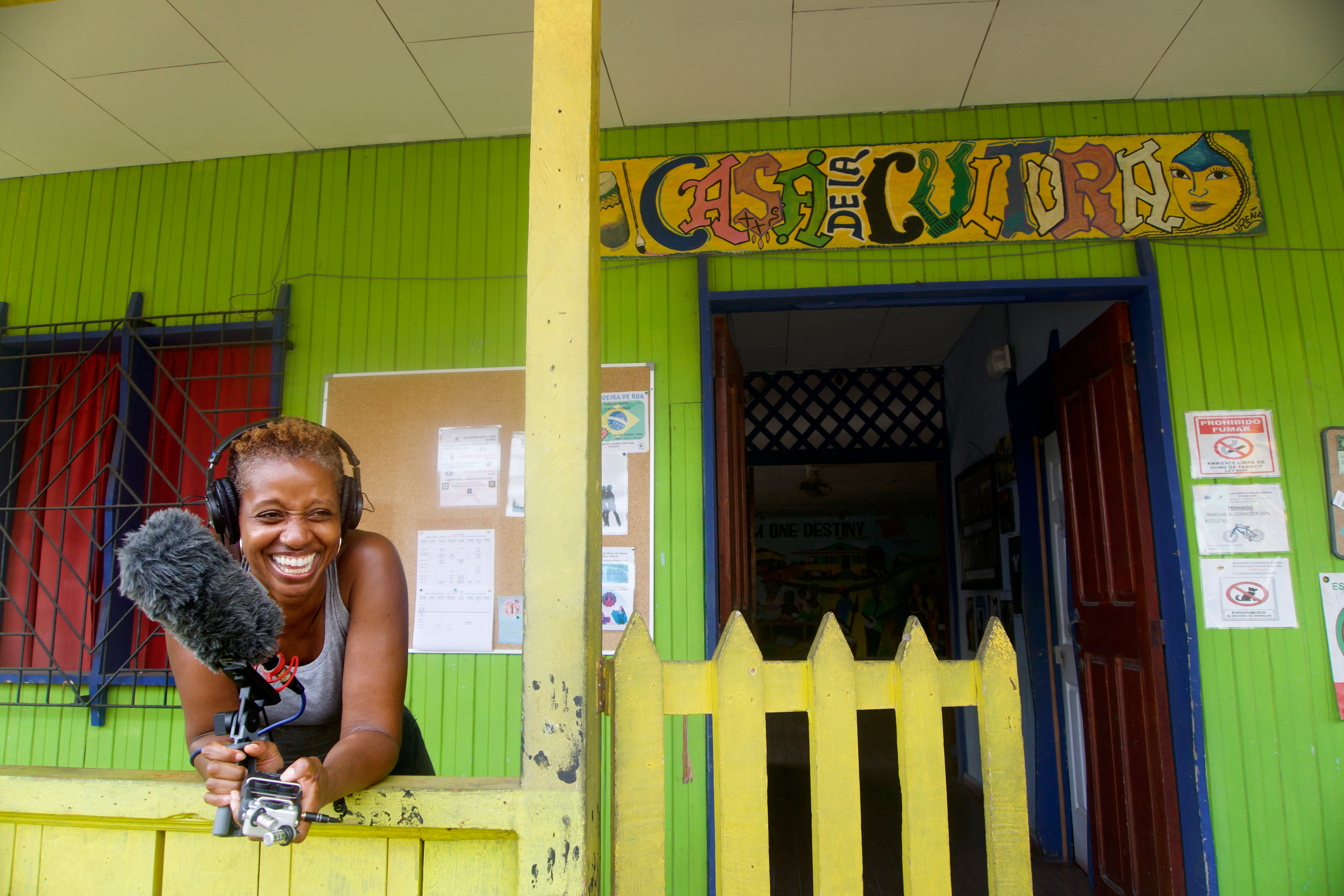 An exterior scene shows a colorful building. Above an open door is a hand-painted sign reading Casa De La Cultura. In front of the door is a simple wooden picket gate. Next to that, a young black woman holds a large microphone and recording device. She is leaning on a railing. She is wearing a large set of headphones. She is smiling as if in the middle of a laugh. 