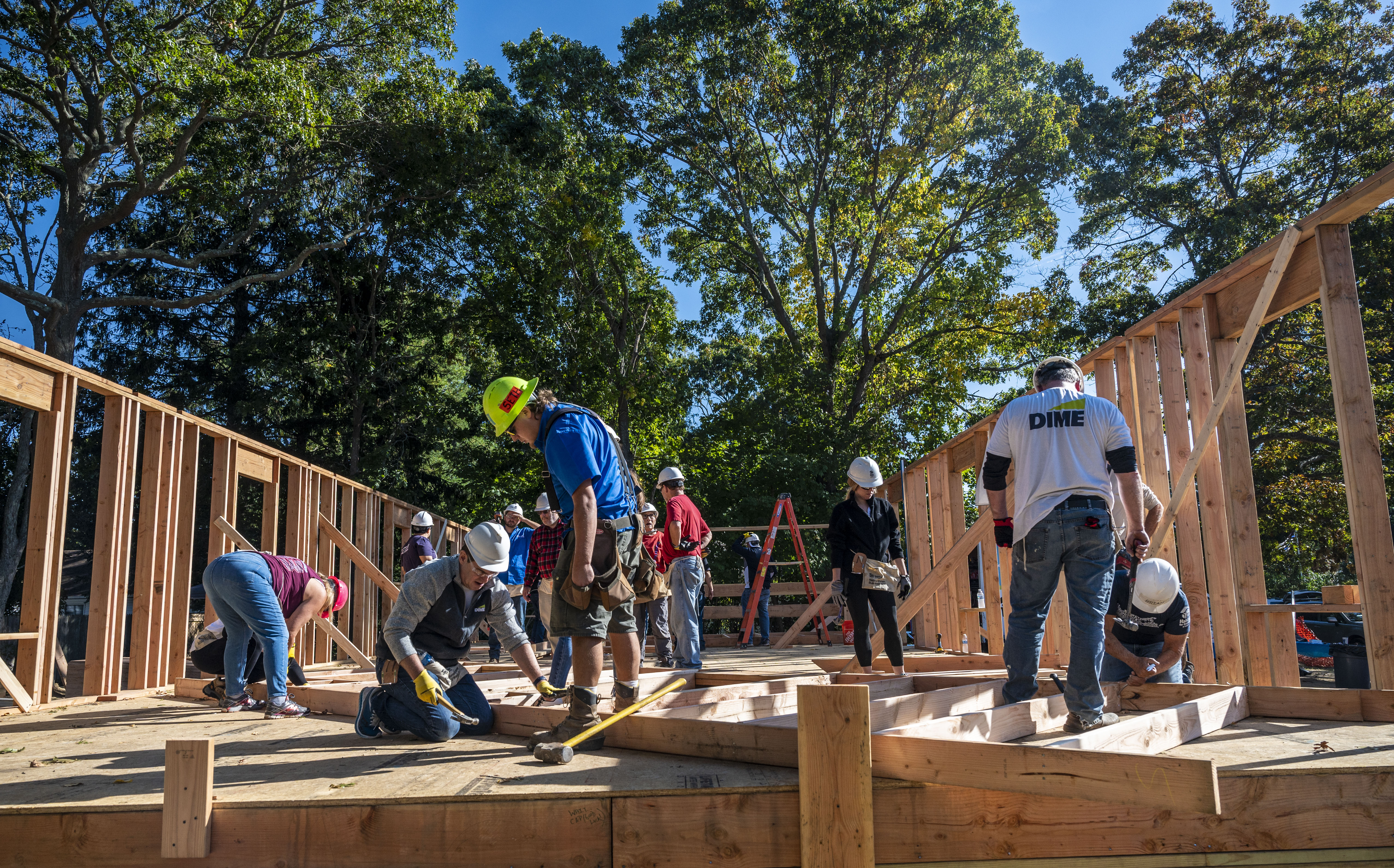 A dozen people in construction worker gear build a home. Only the frame is so-far constructed. 
