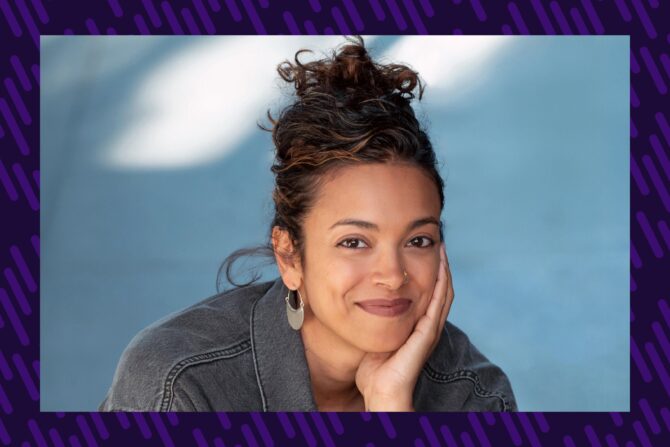 A woman with dark hair smiles at the camera as she rests her chin on her hand.