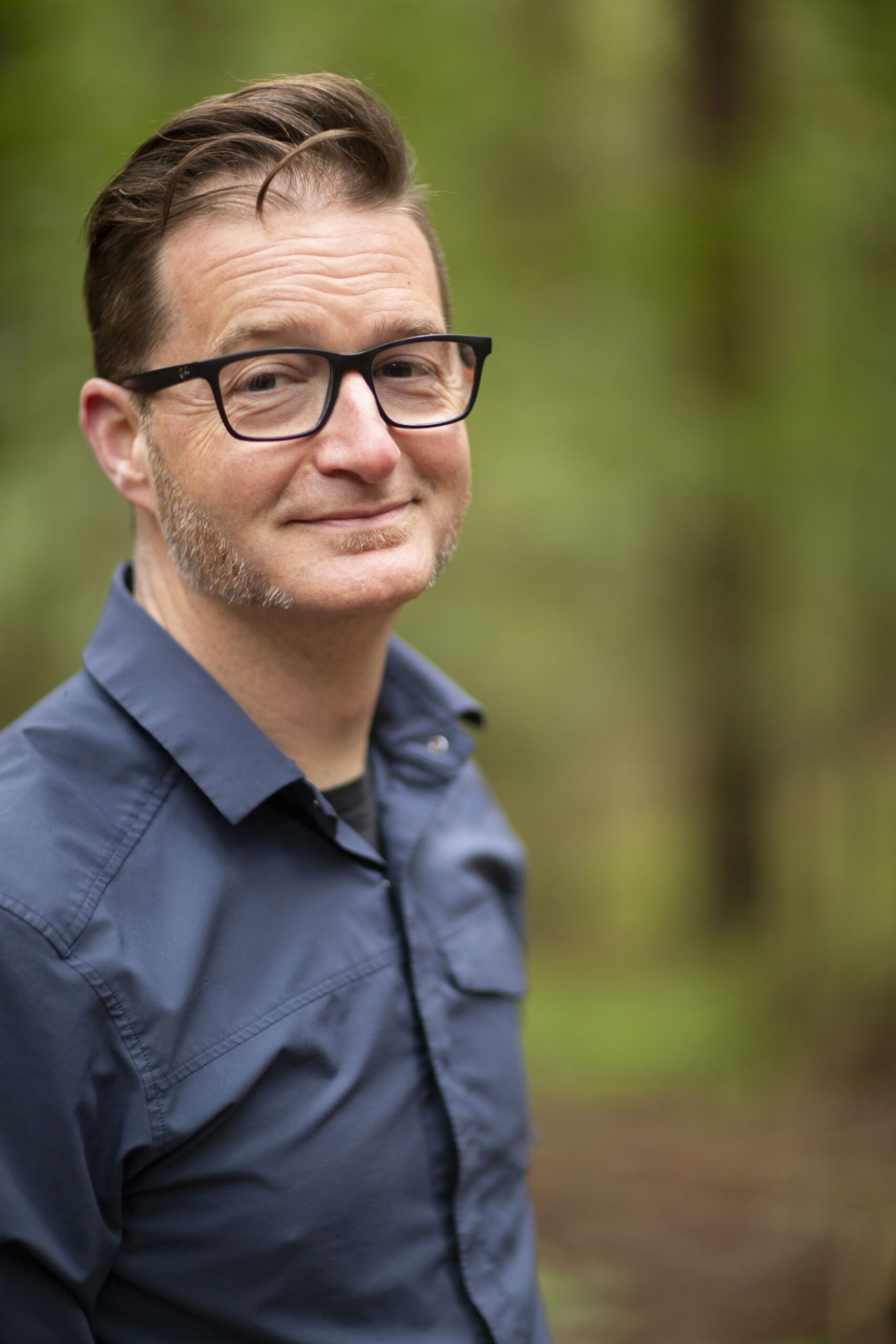 A white man wearing dark-rimmed glasses and a dark-colored button-up shirt stares into the camera with close-lipped smile on his face. He is standing outside against a backdrop of green vegetation.