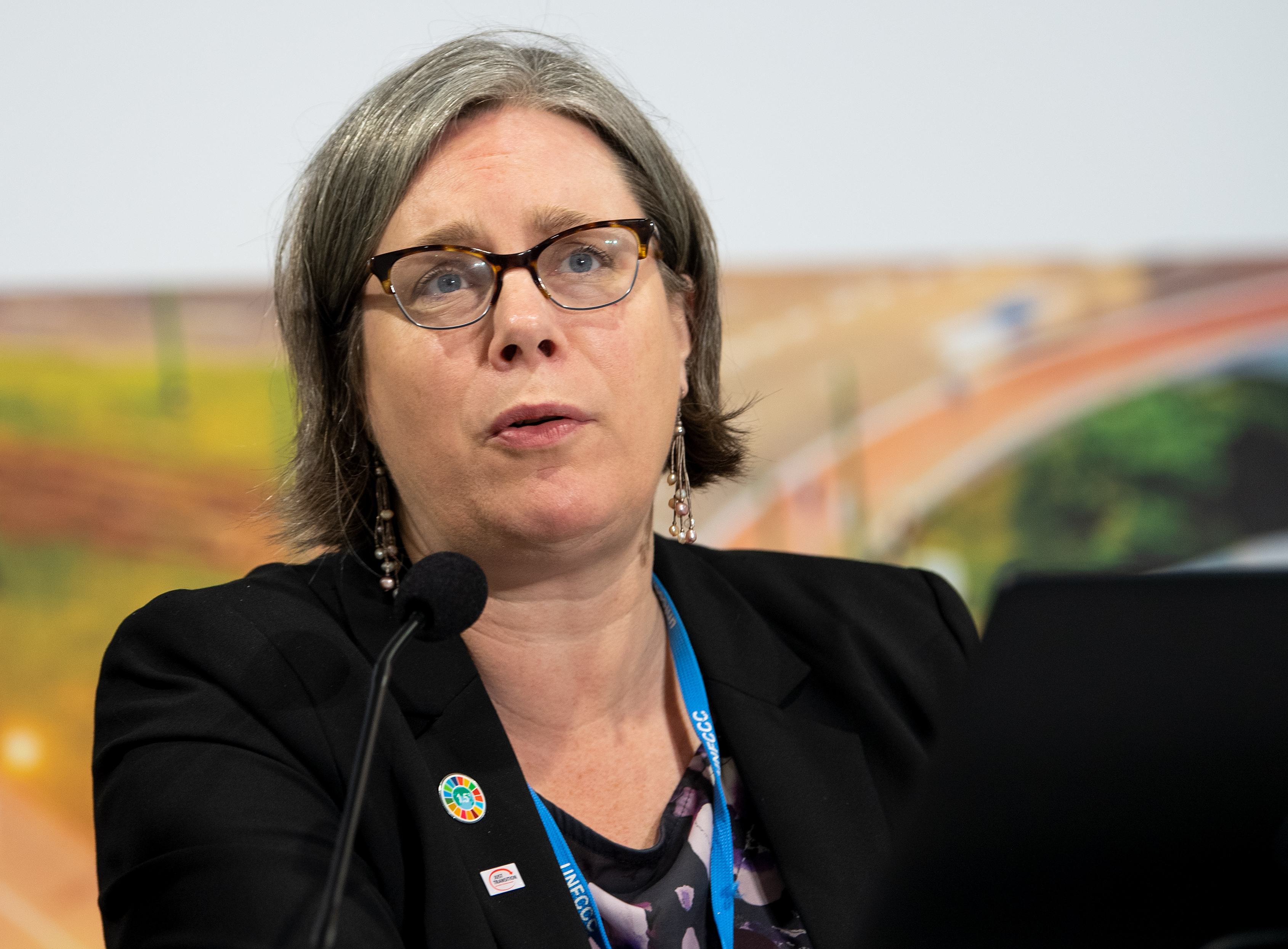 A middle-aged white woman is seen speaking before a small microphone. She is wearing business attire and a lanyard with portions of text visible. The text reads UNFCCC.
