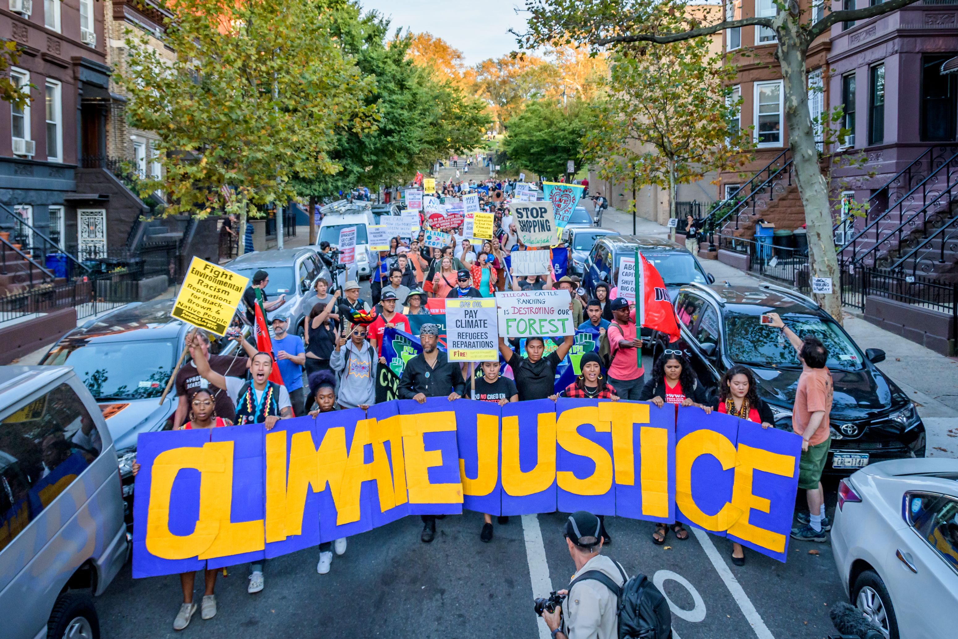 A large crowd of protestors, many holding signs, is seen walking down a residential street in a dense urban area. At the vanguard of the crowd is a large banner reading “Climate Justice.”