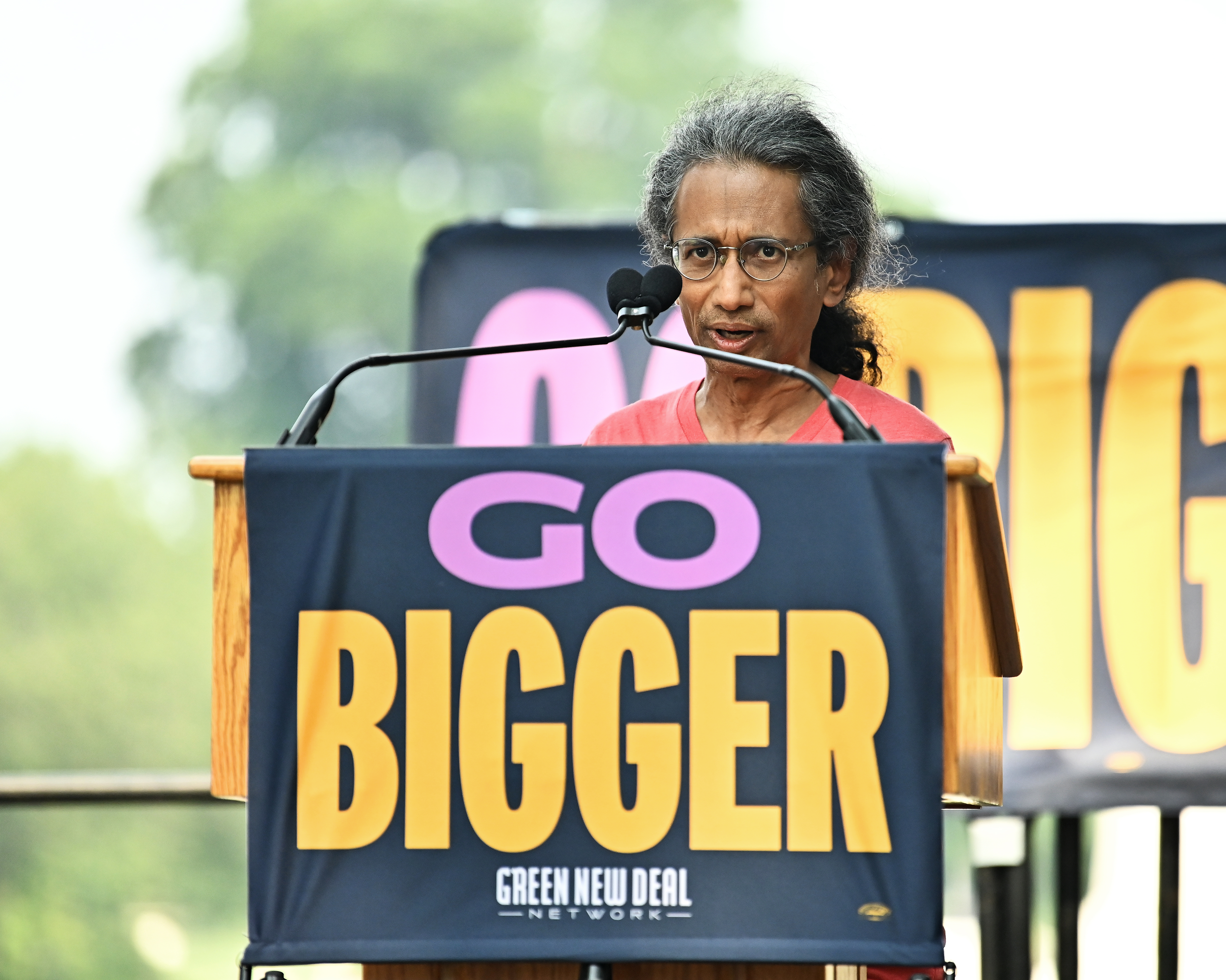 A brown-skinned man with long salt-and-pepper hair pulled back into a ponytail is seen speaking at a podium with two microphones. On the front of the podium is a sign saying “Go Bigger” and in smaller text below that, “Green New Deal.”