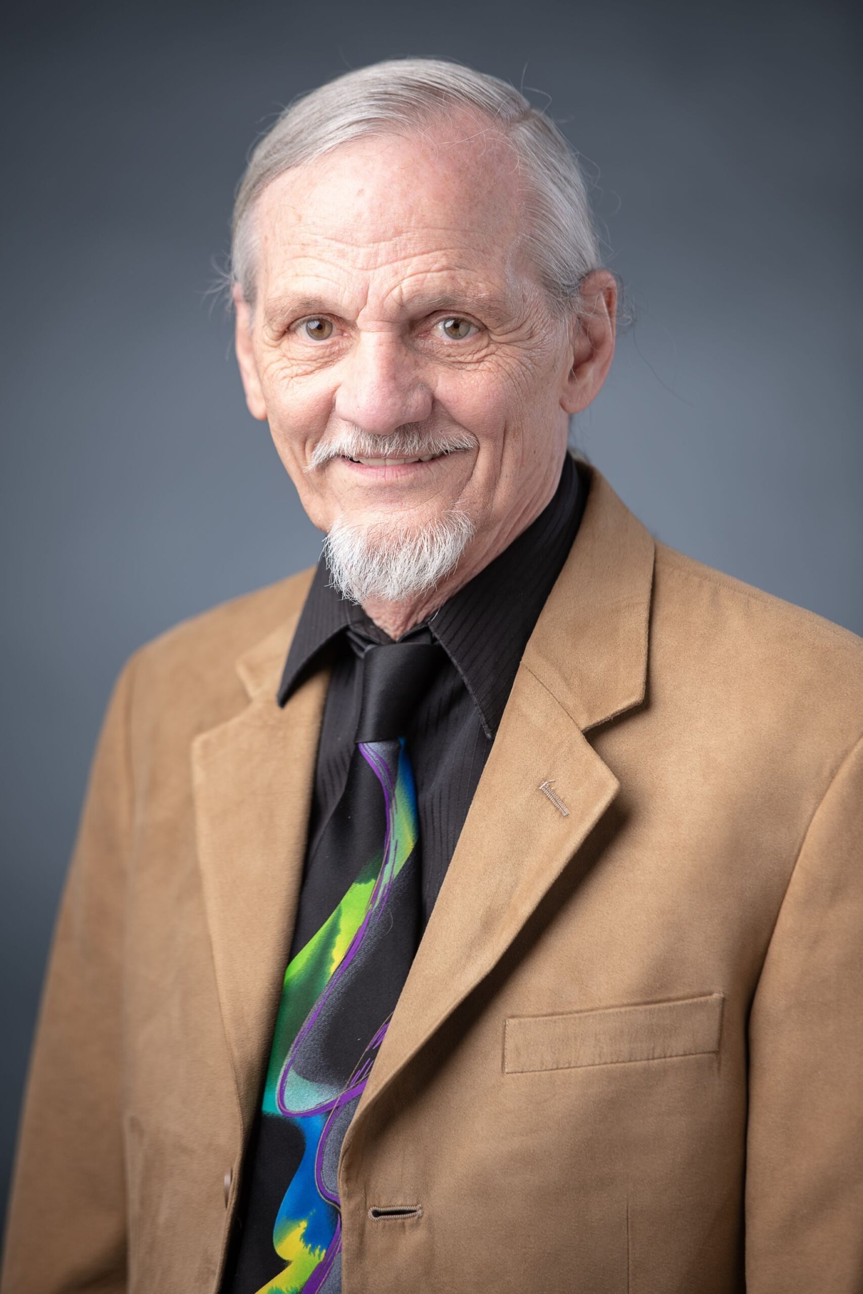 An elderly white man poses for a portrait. He has a white goatee and long white hair. He is wearing a tan jacket, a dark shirt, and a brightly-colored tie. 
