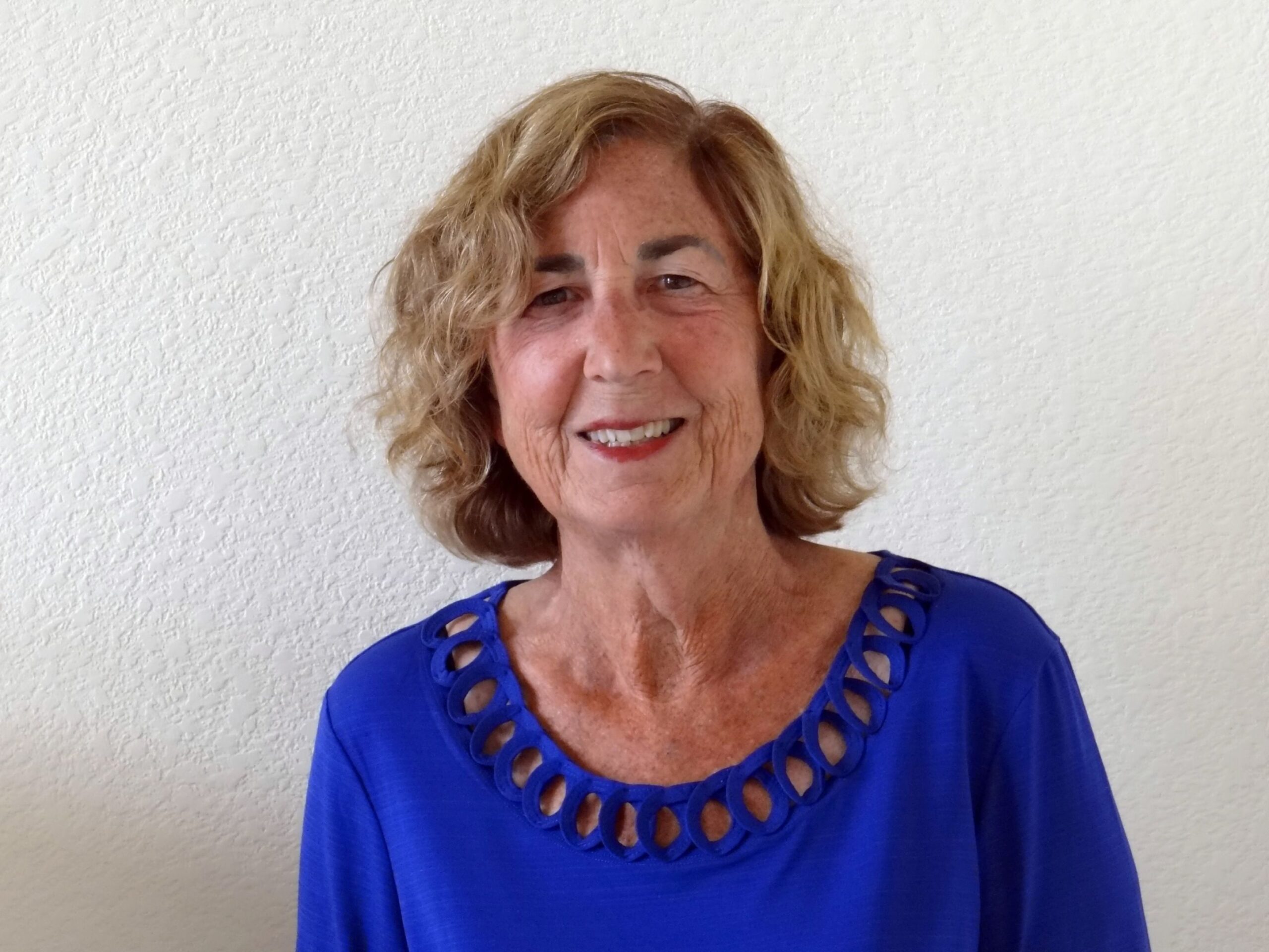 An older white woman smiles for a portrait taken infront of a white wall. She has curly red shoulder-length hair and is wearing a blue blouse with a unique braided neckline.