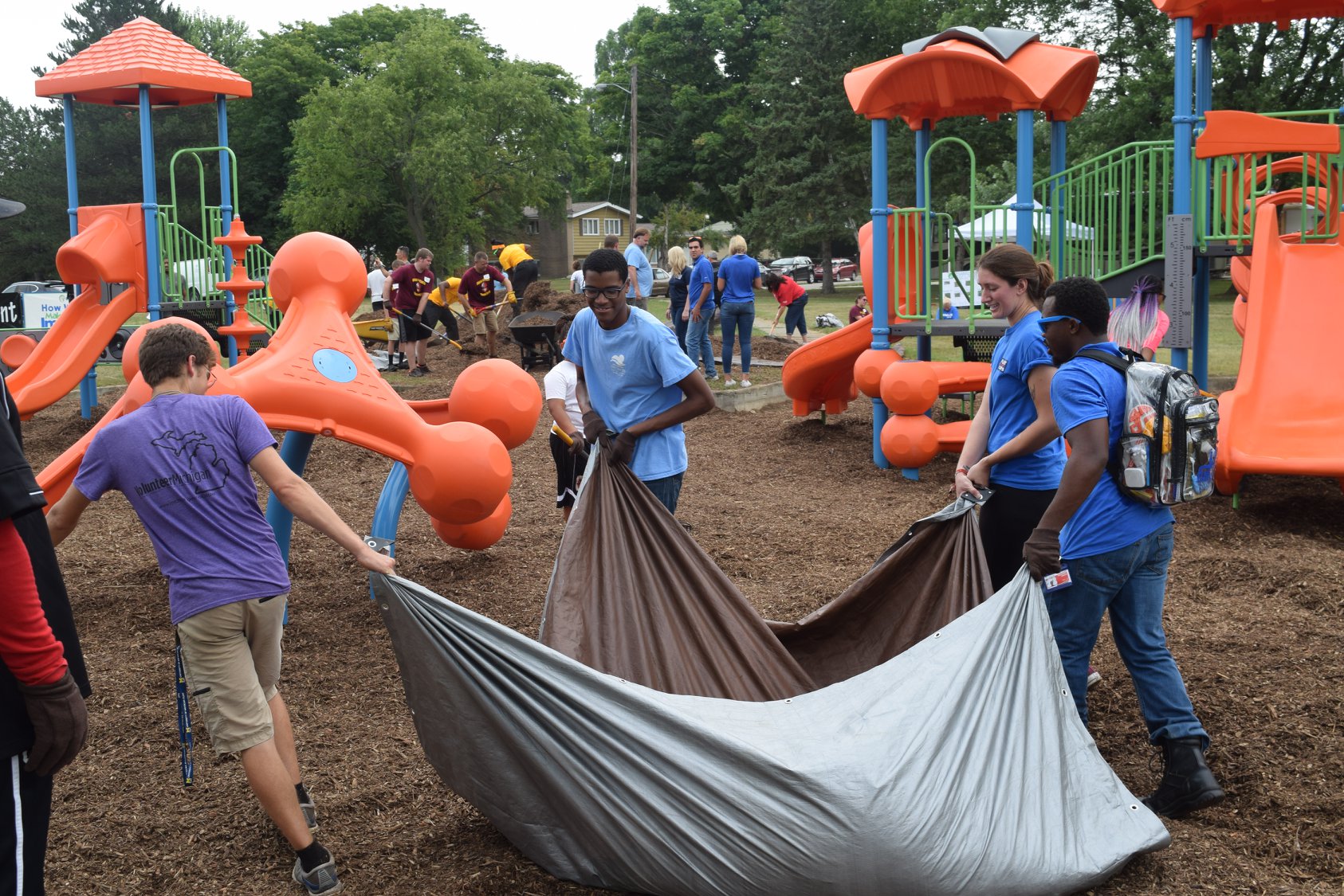 A group of five people drag a large tarp filled with mulch across a new playground. A crowd of people are in the background with shovels. It appears as if they are building or fixing the playground. 
