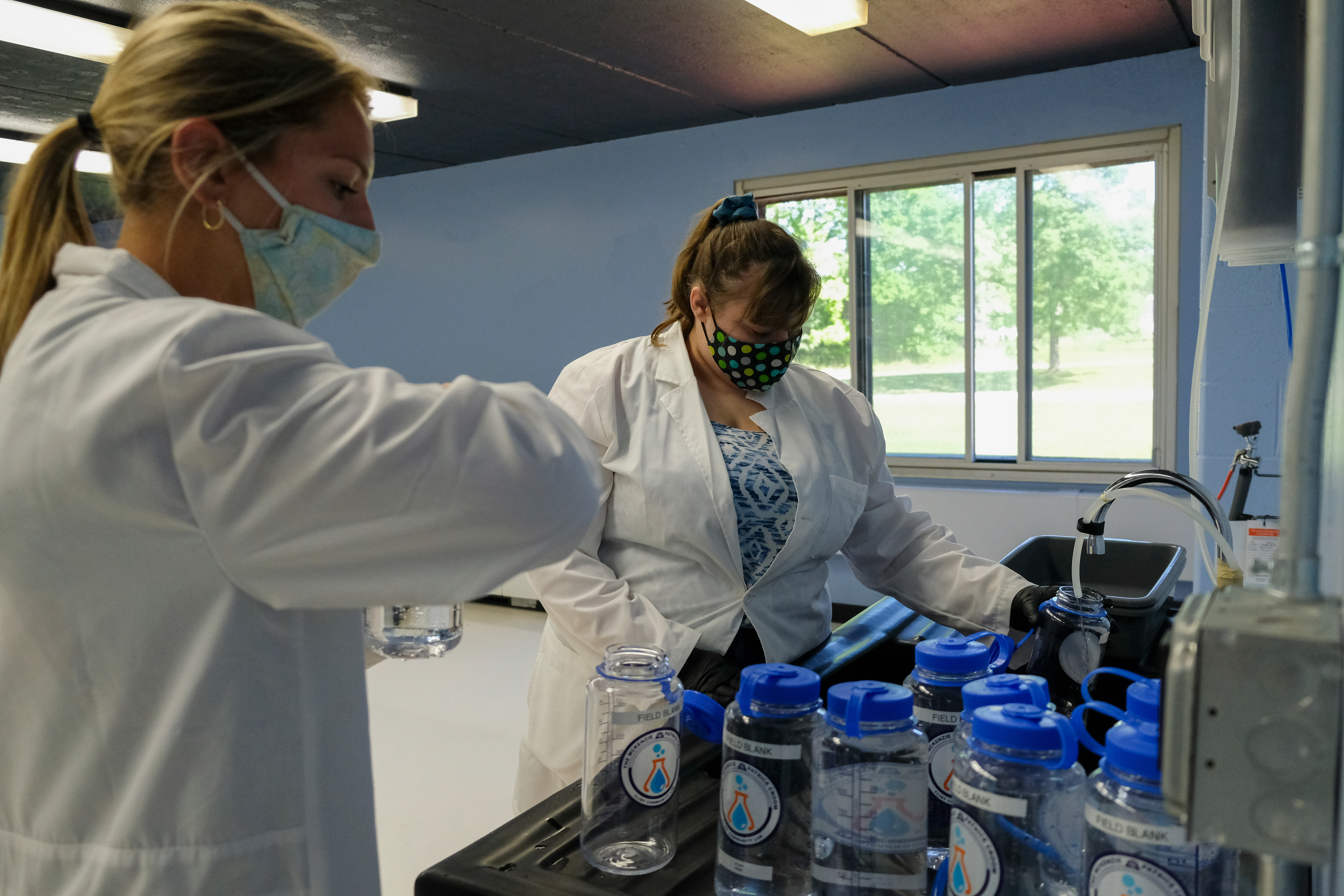 Two white women in lab coats stand next to a sink in a laboratory. 