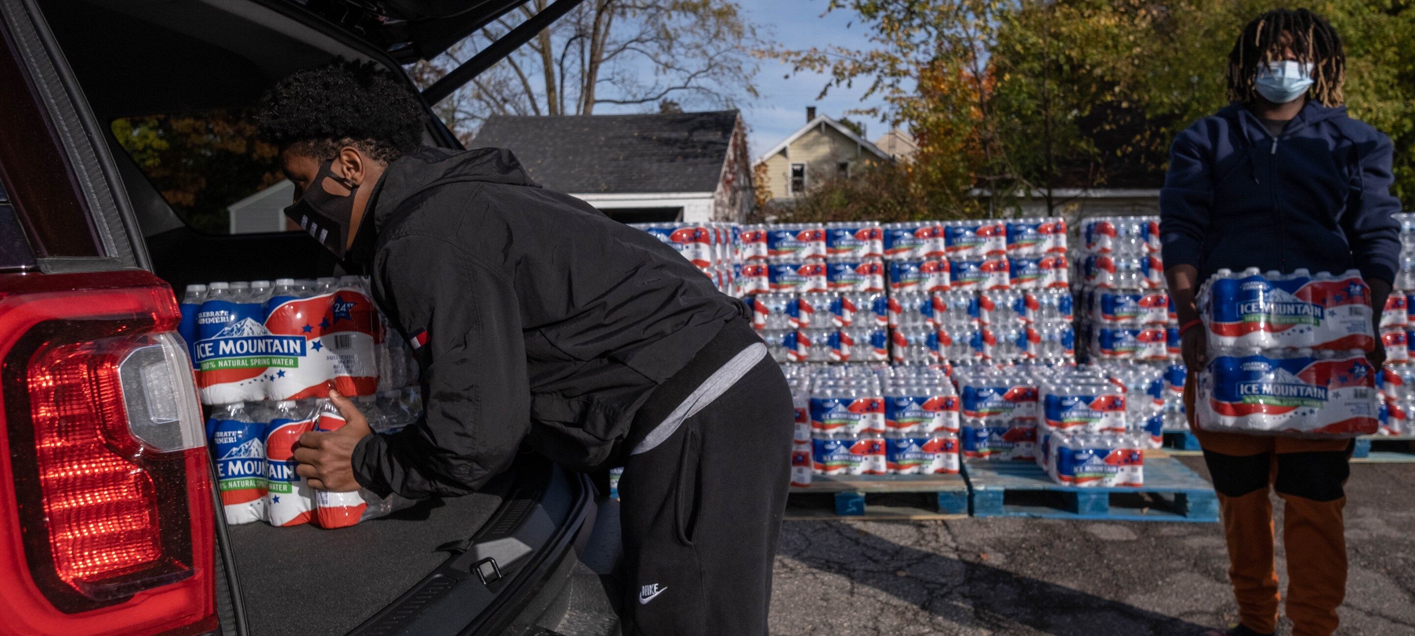 Two black men wearing light jackets and face masks are seen loading pallets of bottled water into the back of a truck.