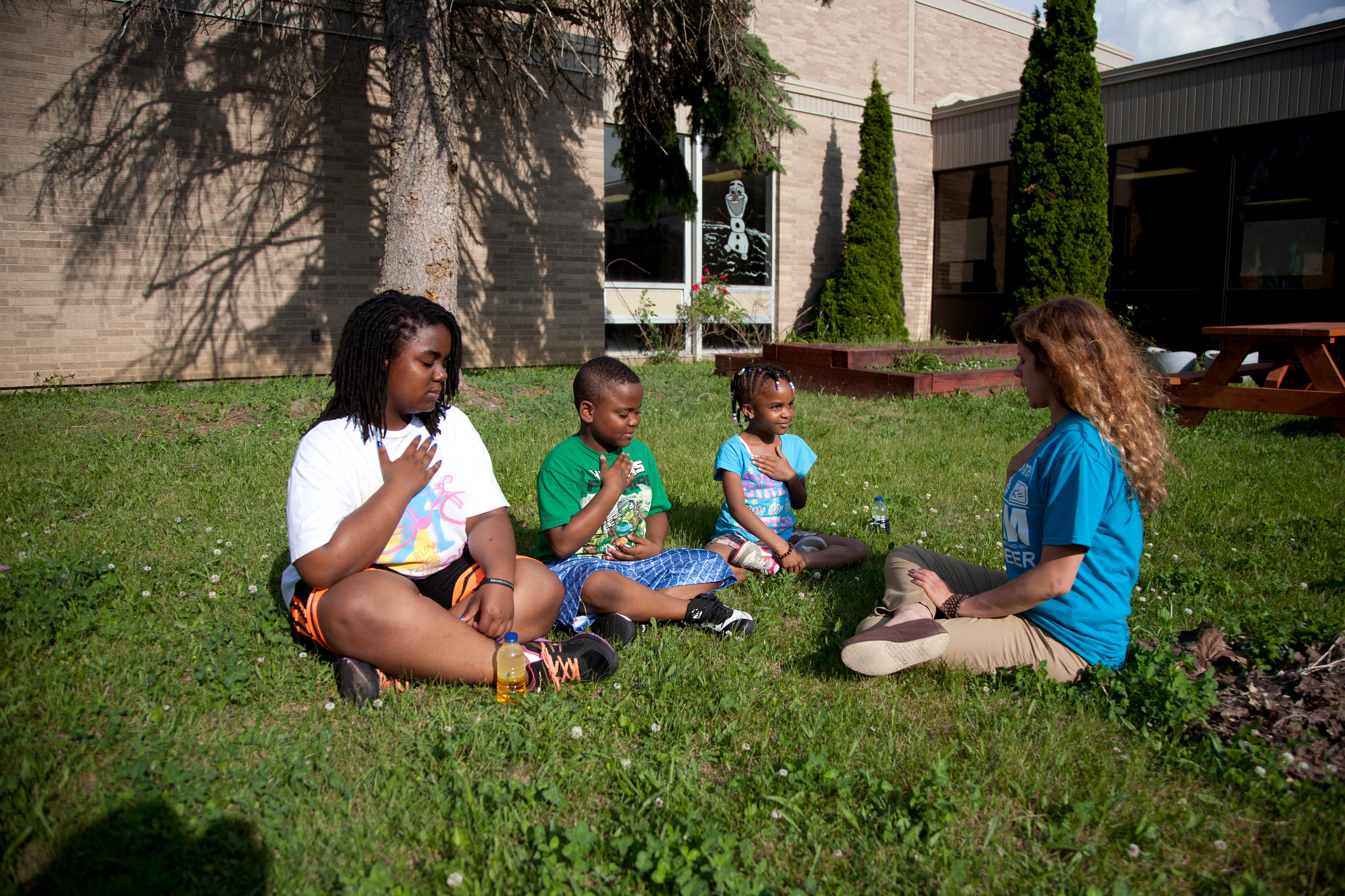 Seated outside, cross-legged, three young black children – two girls and one boy – appear to be meditating under the guidance of of a white female teacher. 