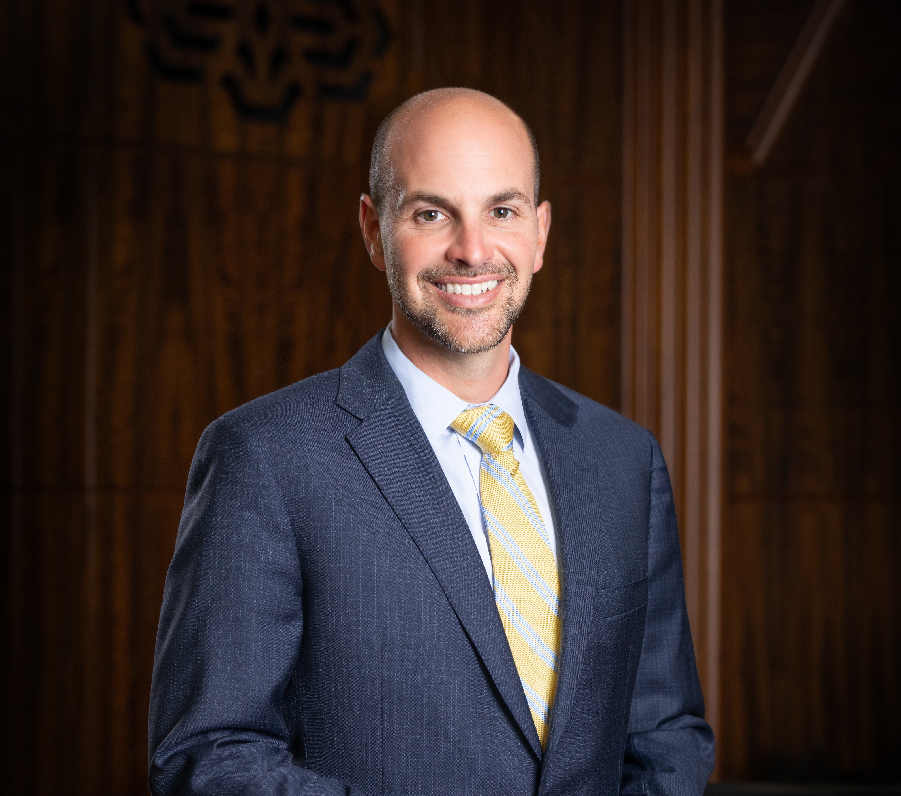 A white man, bald, with a short beard, is seen smiling for a portrait. He is wearing a suit and tie. 