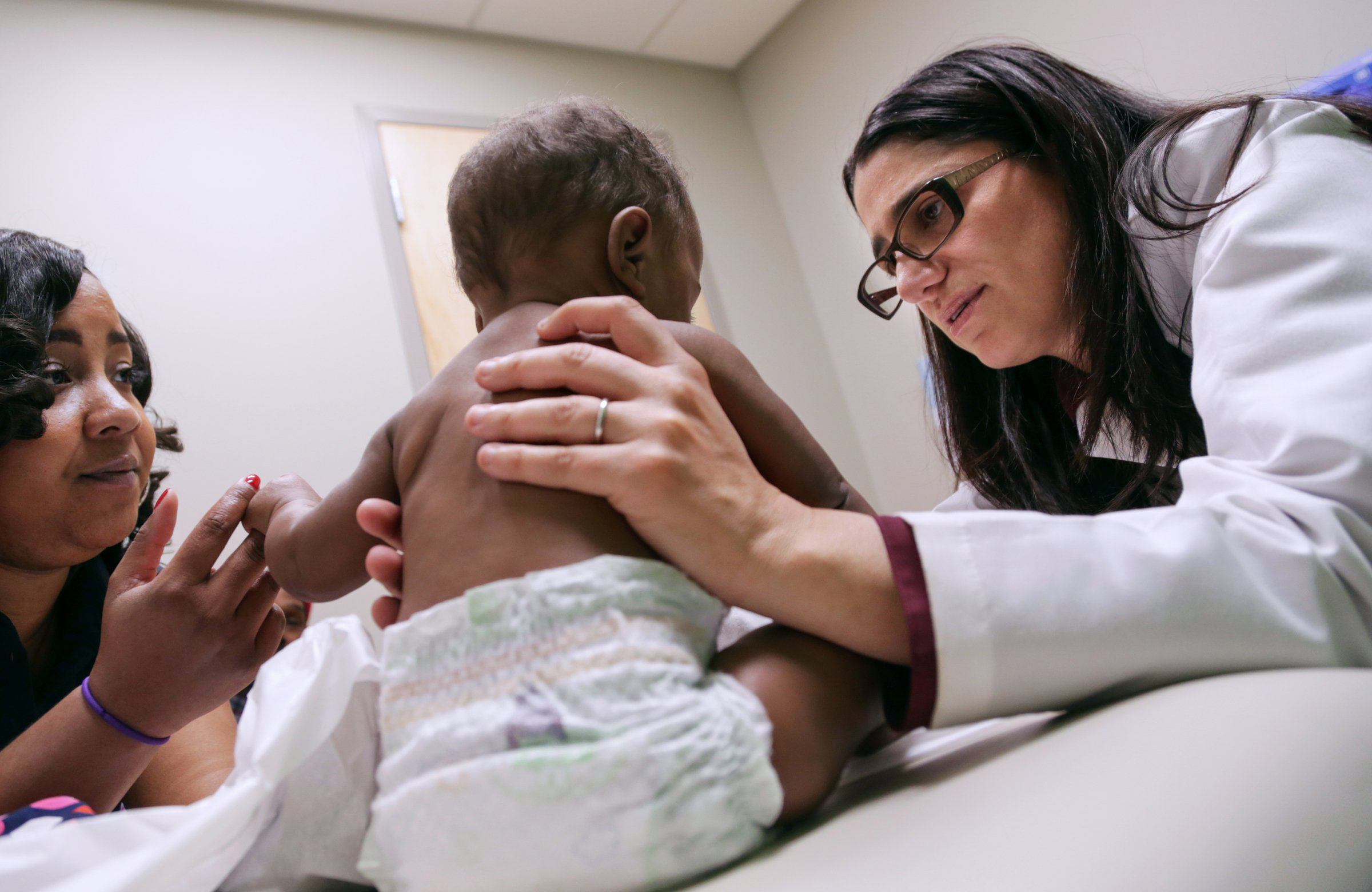 A female doctor, white skin, brown hair, with square-framed glasses examines a very-young brown-skinned infant. The baby is holding her mother’s hand. Her mother is seen, also brown-skinned. 