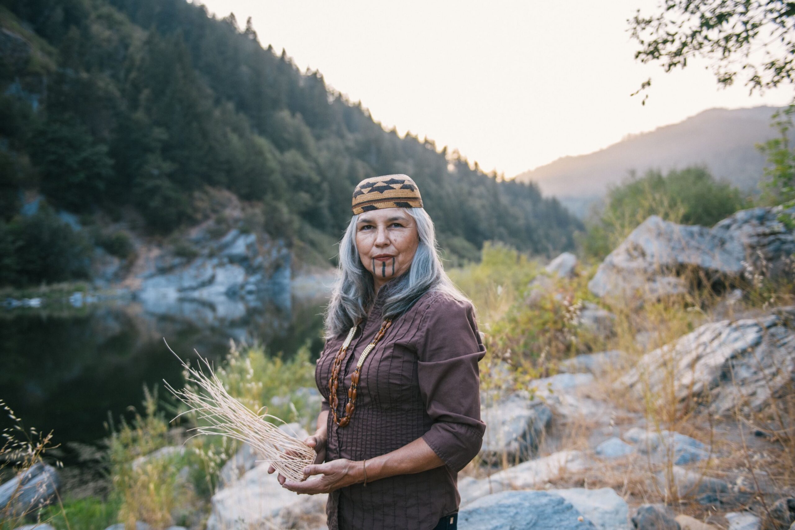 Portrait of Margo Robbins in Weitchpec, Calif. on August 23, 2021 in front of the Klamath River, holding a basket she is in the process of making from fire-dependent resources.