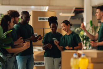 A group of volunteers wearing matching green shirts stand together smiling and looking at a phone, surrounded by large boxes labeled ‘Donation’ in a bright, indoor workspace.