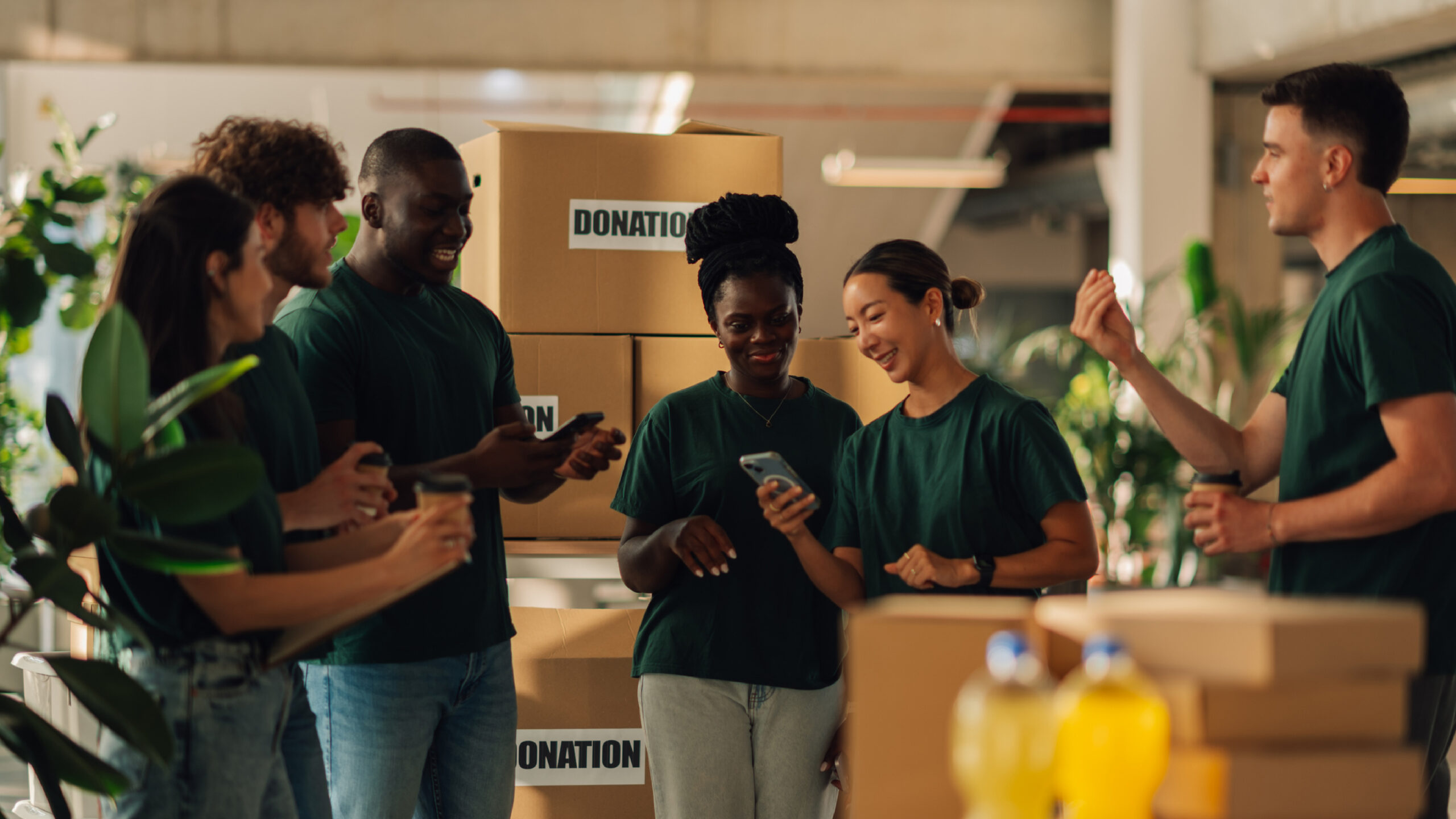 A group of volunteers wearing matching green shirts stand together smiling and looking at a phone, surrounded by large boxes labeled ‘Donation’ in a bright, indoor workspace.