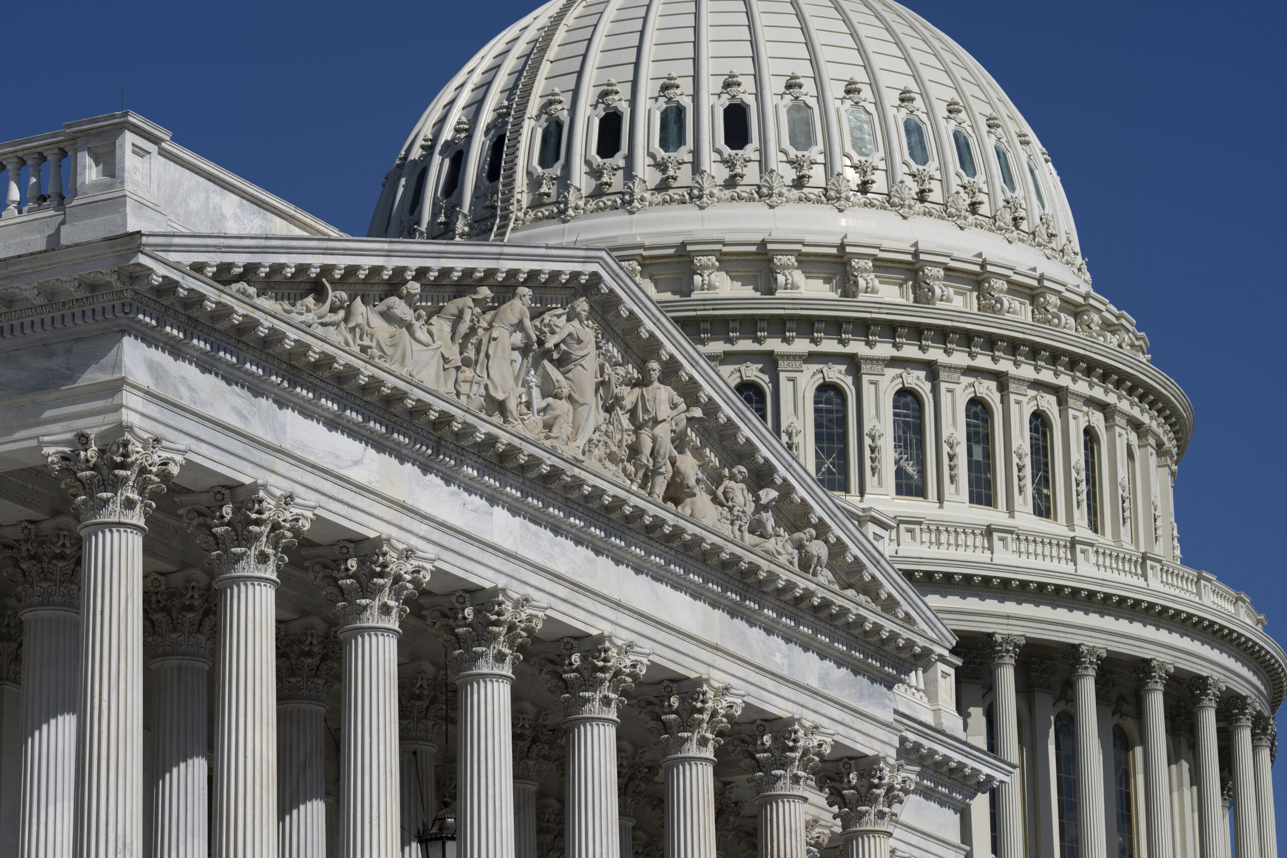 A low-angle view of the white marble U.S. Capitol Building, showing its detailed pediment with sculptures, Corinthian columns, and the ribbed dome against a clear blue sky.