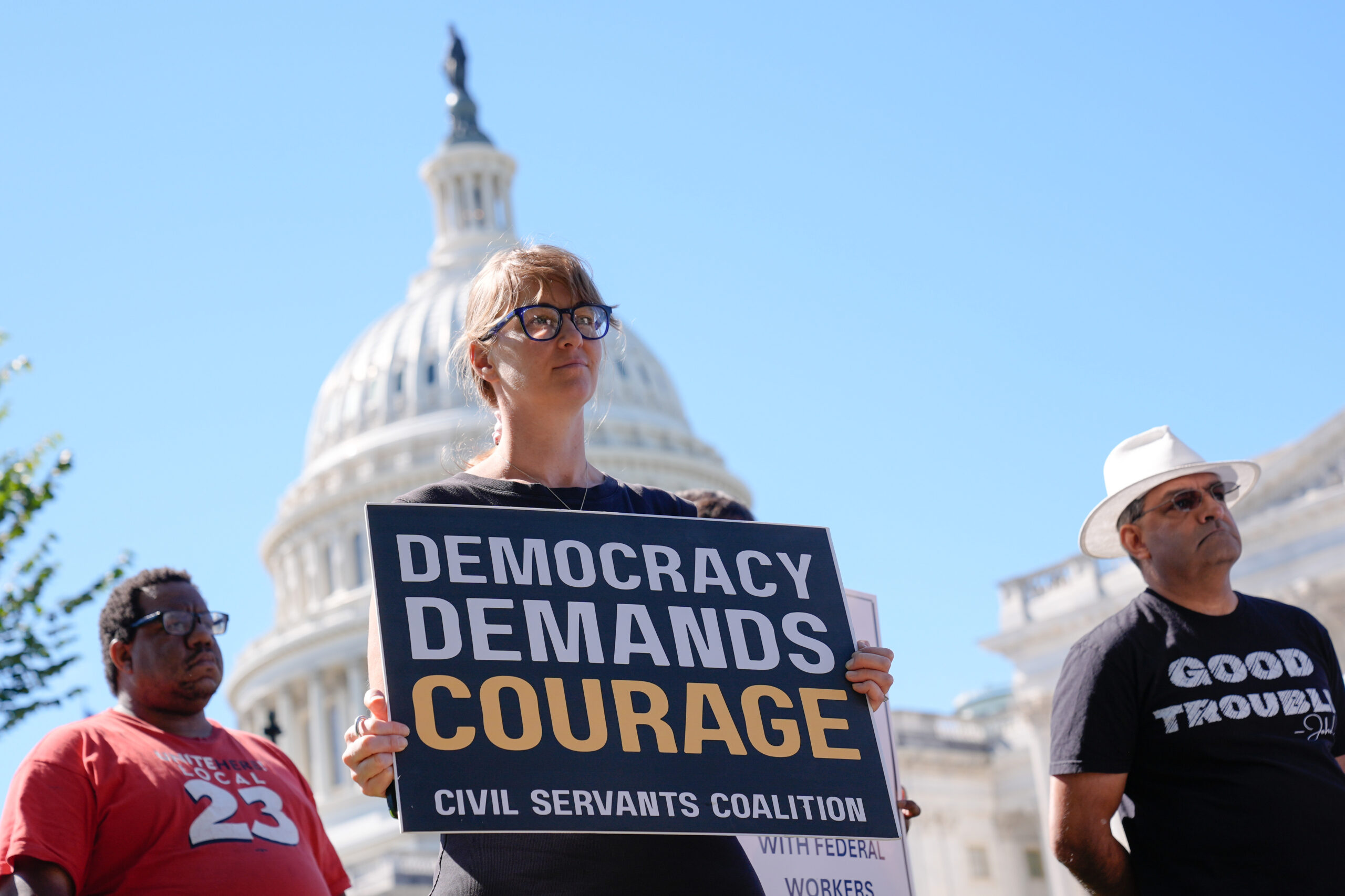 A woman holds a sign reading 'DEMOCRACY DEMANDS COURAGE' with two men beside her, all looking towards the U.S. Capitol Building under a clear blue sky.