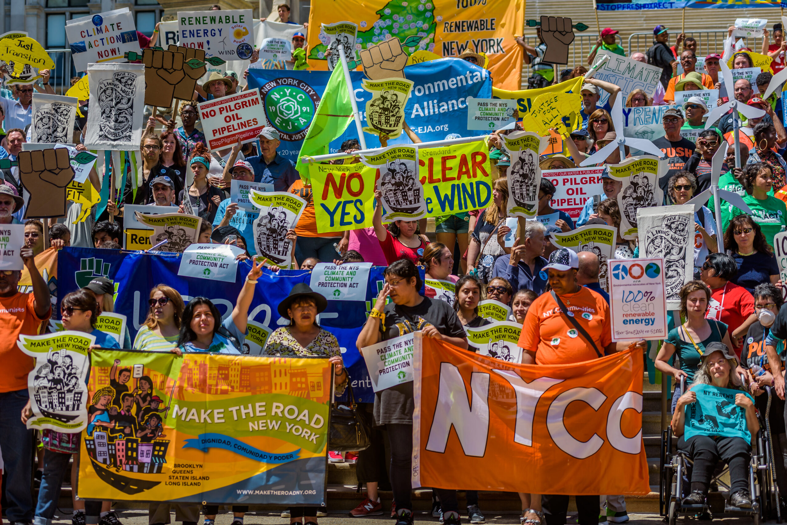 A large crowd of diverse people at an outdoor protest rally, holding numerous signs and banners advocating for climate action, renewable energy, and environmental justice in New York. Signs read "Climate Action Now," "Renewable Energy Now," "No Pilgrim Oil Pipelines," and "Make the Road New York." Some signs feature raised fists and wind turbine cutouts.