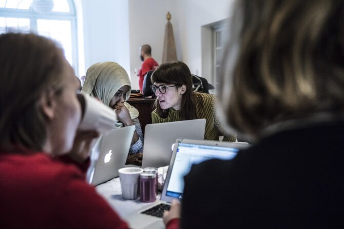 Four people, two in focus, work on laptops at a table. A woman in a hijab and another in glasses intently look at their screens or each other, suggesting collaboration. Bright light streams from a large window in the background.