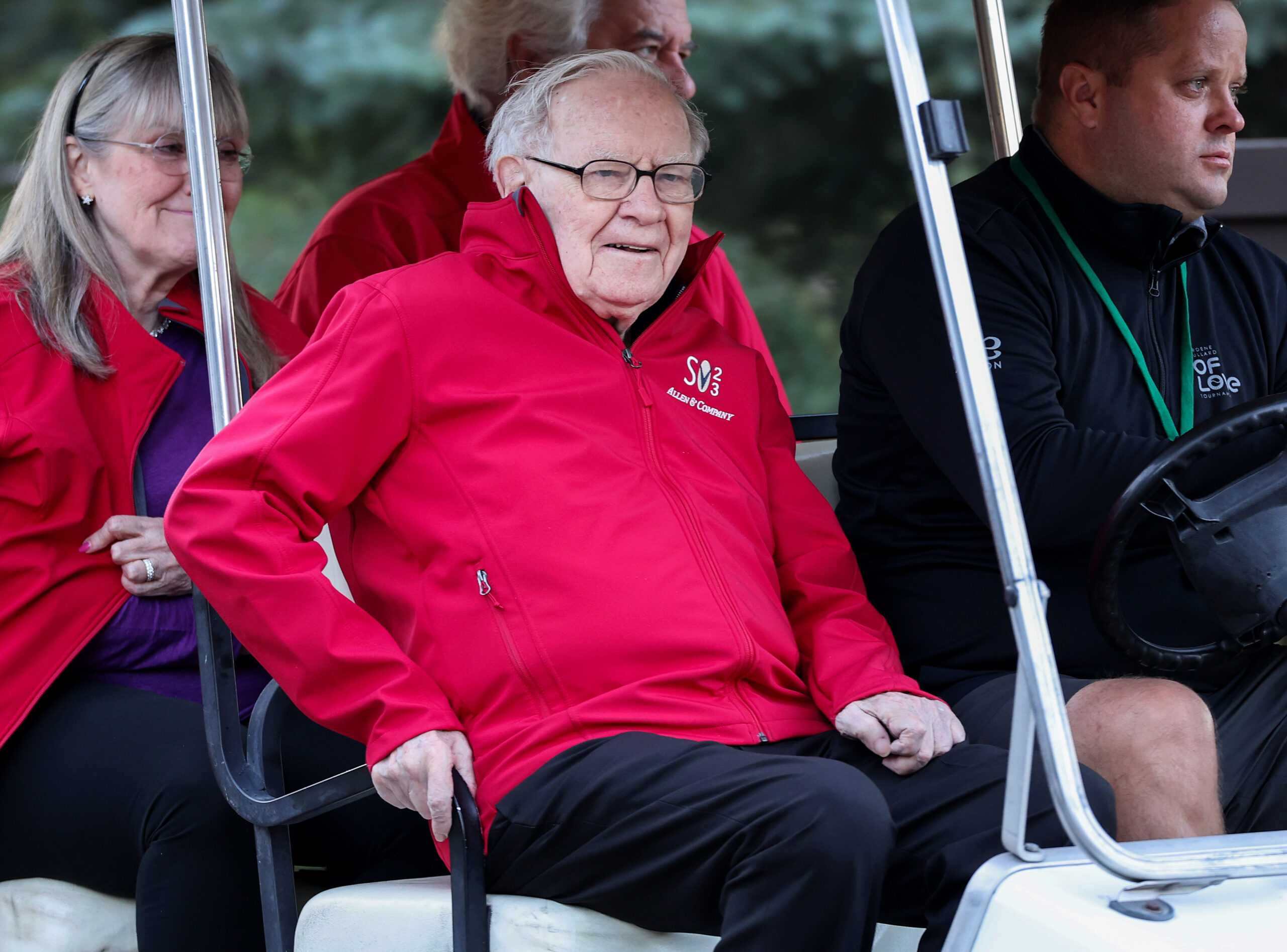 Warren Buffett, an elderly man with white hair and glasses, smiles while sitting in a golf cart, wearing a red jacket with 'Allen & Company' logo. Other people are partially visible around him.