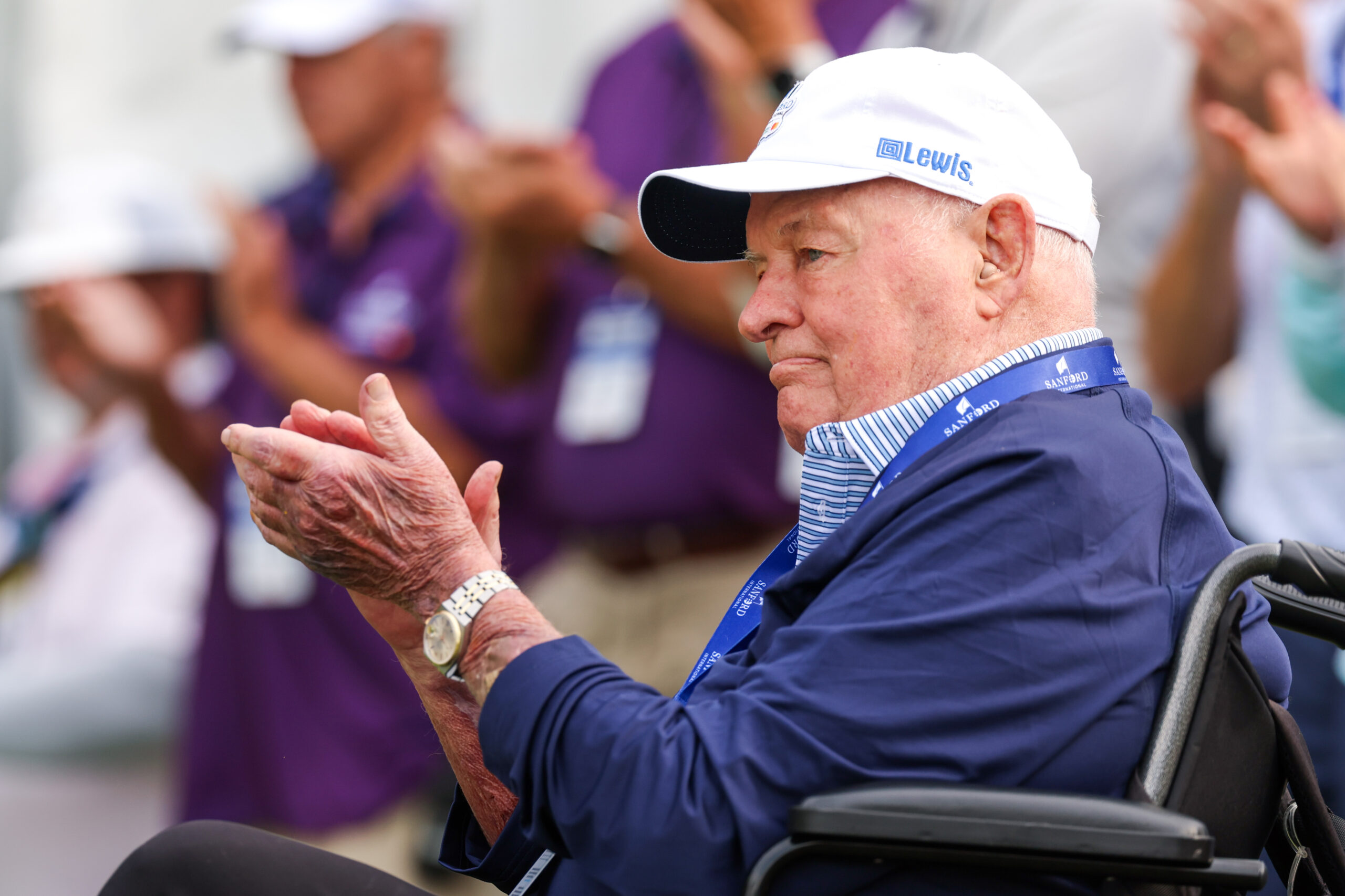 An elderly man in a white baseball cap, blue jacket, and a 'Sanford International' lanyard claps his hands while seated, looking to the right.