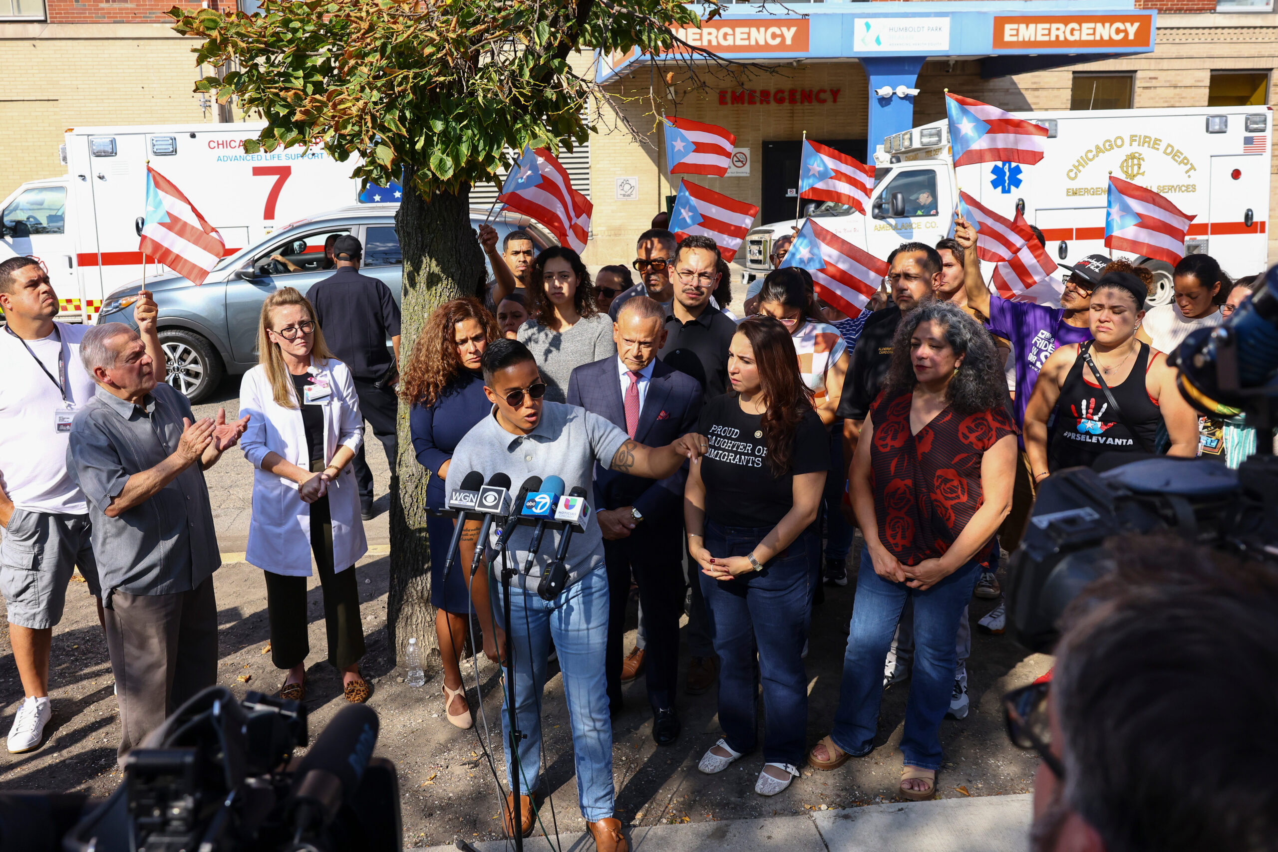 A diverse group of people attend an outdoor press conference in front of ambulances and an emergency building. A speaker addresses microphones.