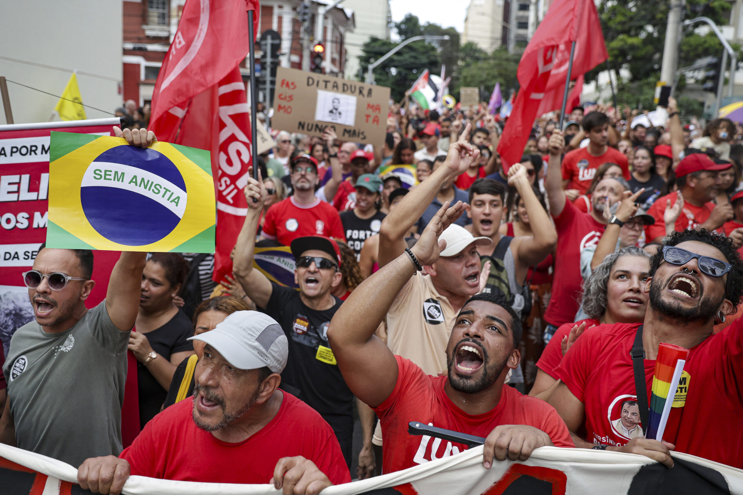 A large, diverse crowd of protesters on a street, many wearing red, shouting and raising fists. A central sign features the Brazilian flag with "SEM ANISTIA" (No Amnesty), while another sign reads "DITADURA NUNCA MAIS" (Dictatorship Never Again). Red flags and a rainbow flag are visible.