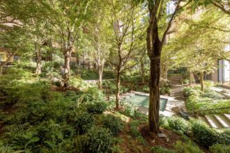 A lush, multi-level urban garden with dense green trees, ferns, and shrubs. Modern buildings are visible through the foliage, and stone stairs with handrails traverse the sloped landscape. A rectangular pond is in the mid-ground, and a person is gardening on the left.