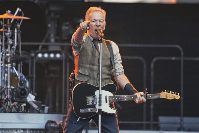 Bruce Springsteen on stage, singing into a microphone, holding a black electric guitar, and pointing forward with his right hand. He wears a waistcoat and tie.