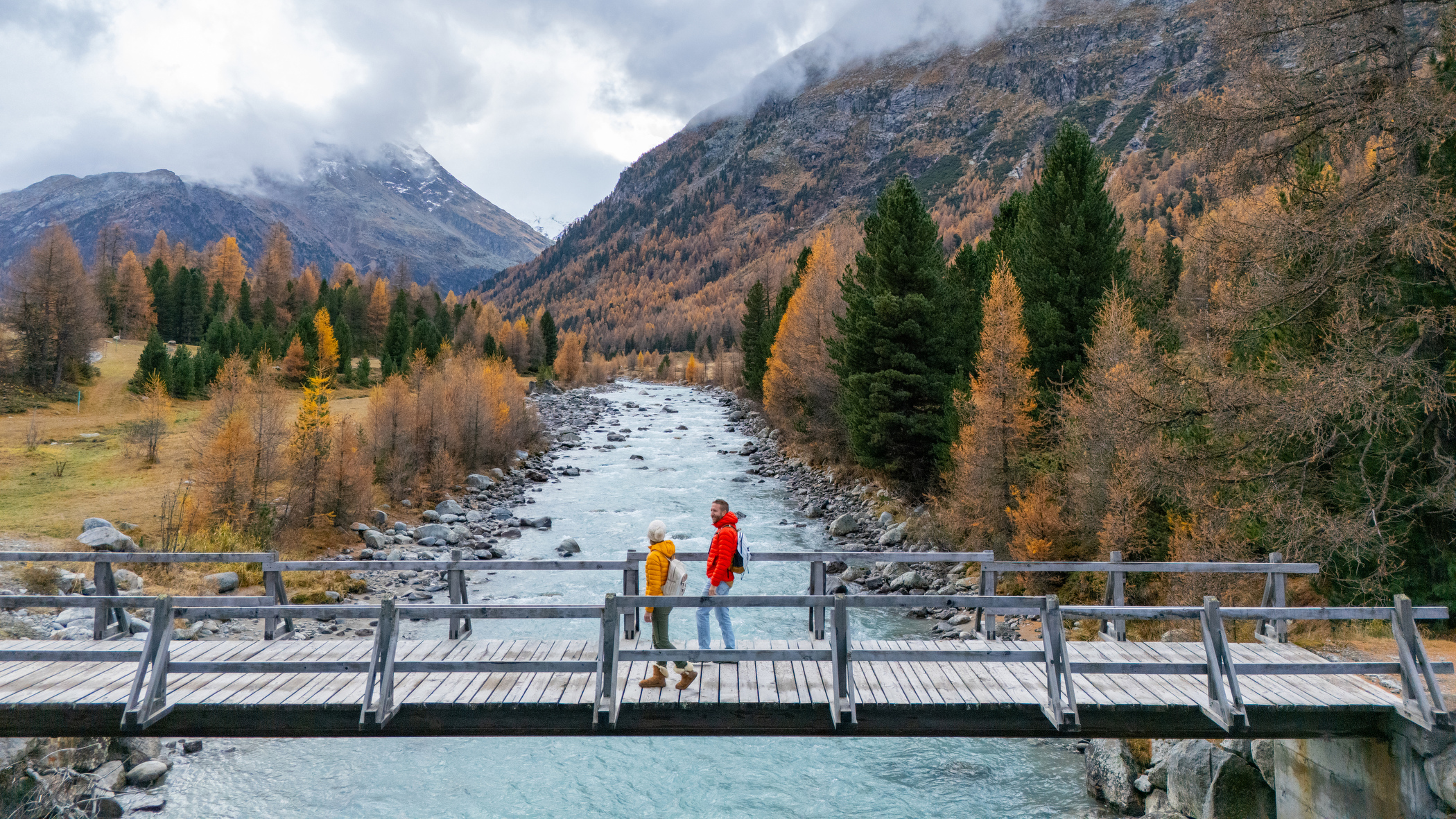 Two people on a wooden bridge over a turquoise river, surrounded by golden autumn trees and cloud-covered mountains.