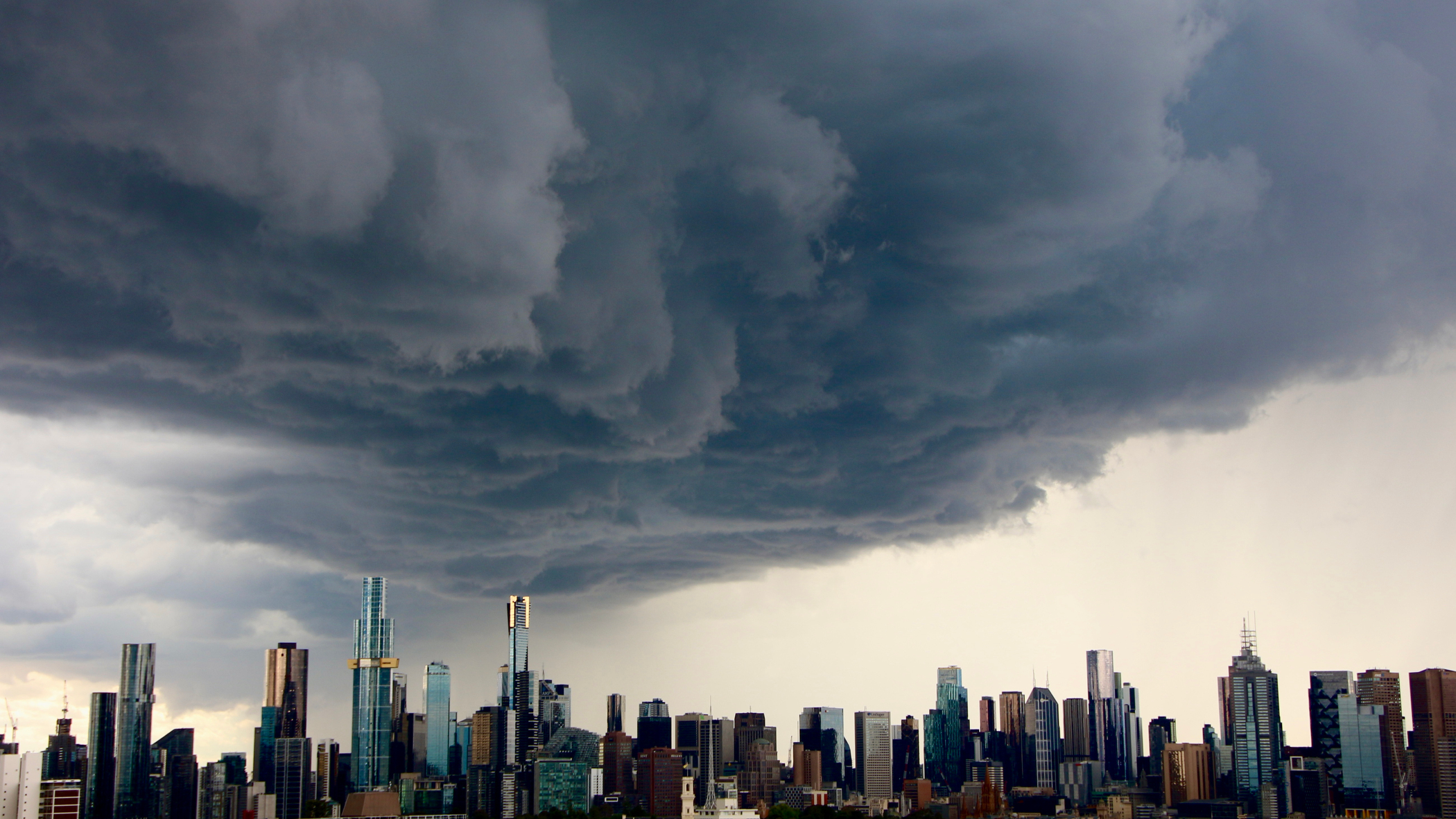A wide shot of a modern city skyline with numerous skyscrapers beneath a vast, dark, and turbulent storm cloud formation.