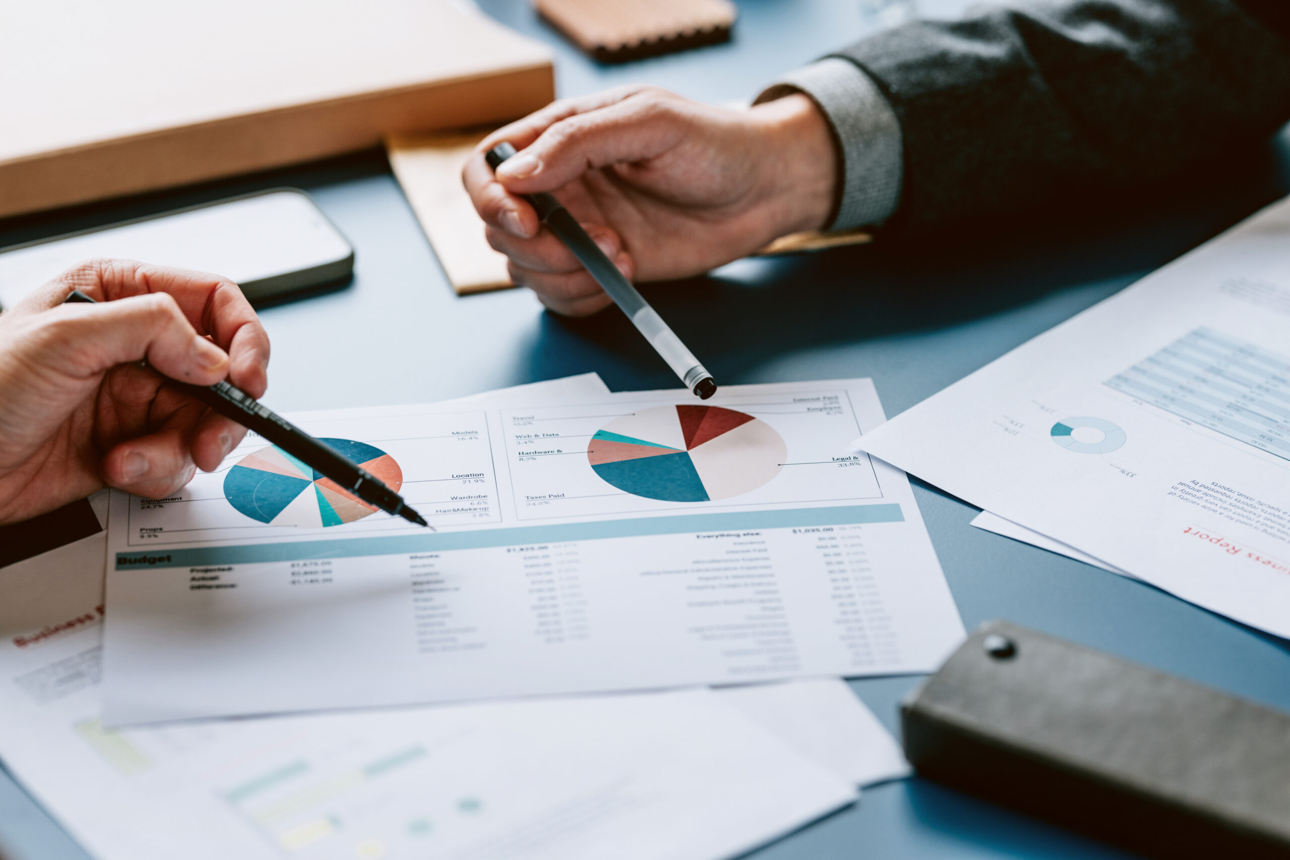 Two people's hands pointing with pens at business documents with pie charts and budget details on a blue table.
