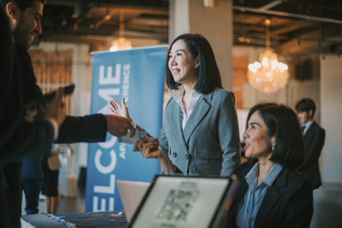 Three people at an event registration desk. A smiling woman in a gray suit receives badges, a man holds a phone, and another woman sits near a laptop displaying a QR code.