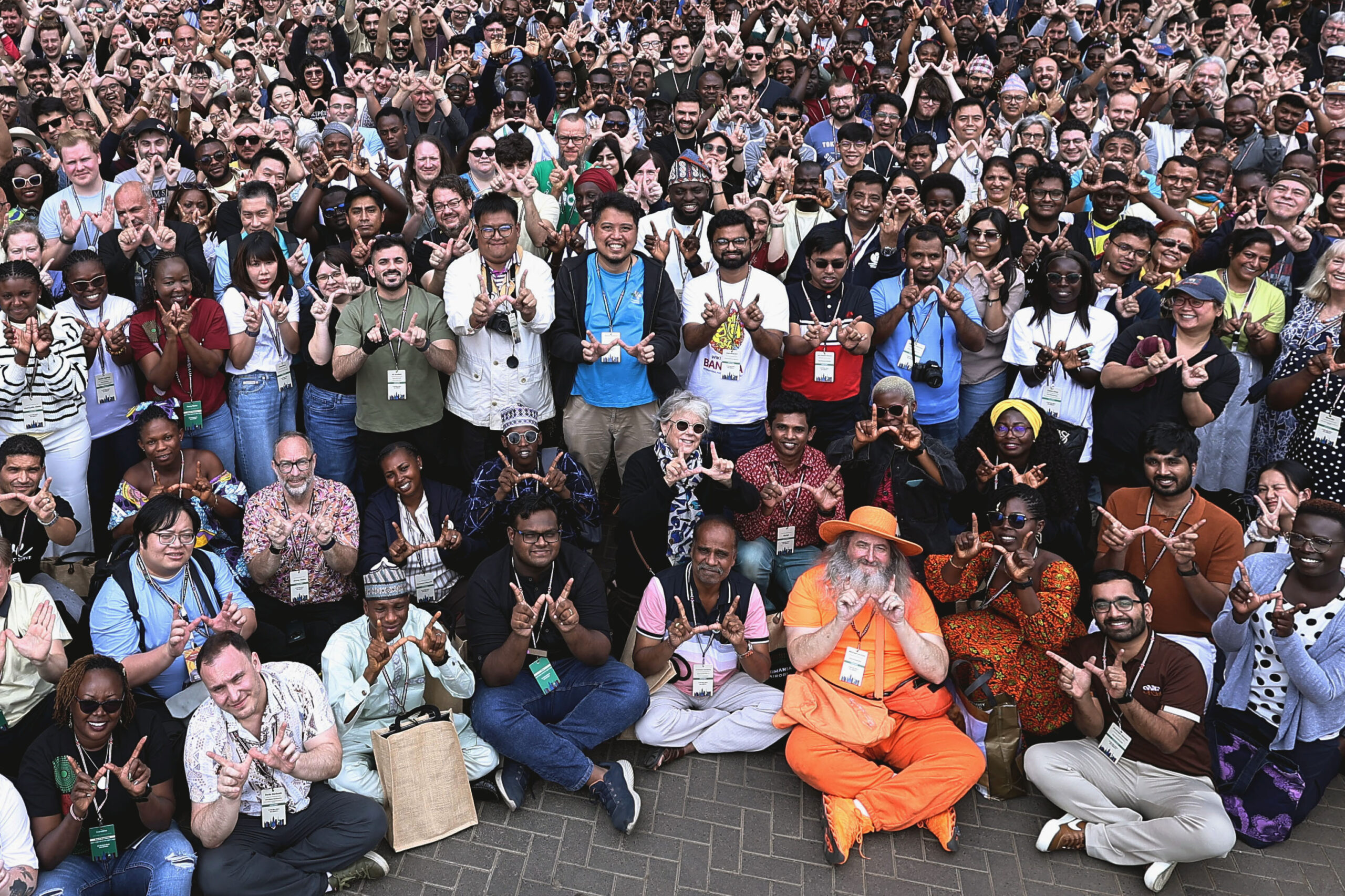 A large, diverse group of people from various backgrounds smiling and making a 'W' hand gesture with their hands, gathered outdoors at a Wikimedia event.