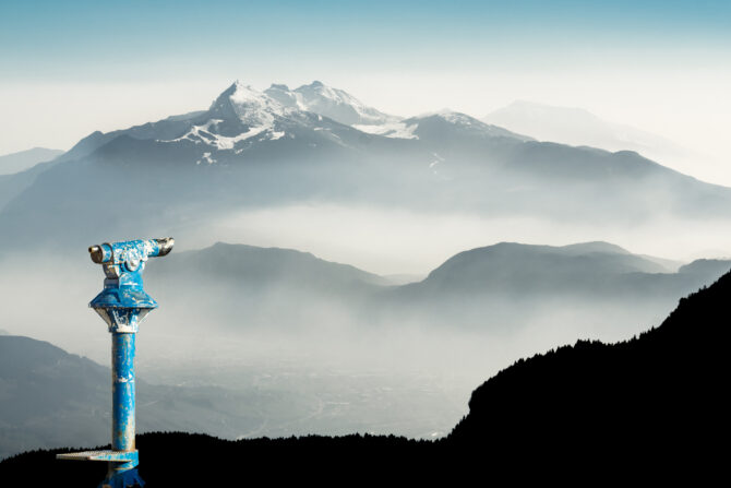 A weathered blue coin-operated telescope stands in the foreground, overlooking a vast, layered mountain range partially obscured by mist. Distant snow-capped peaks rise under a pale blue sky, with a dark forested silhouette framing the bottom right.