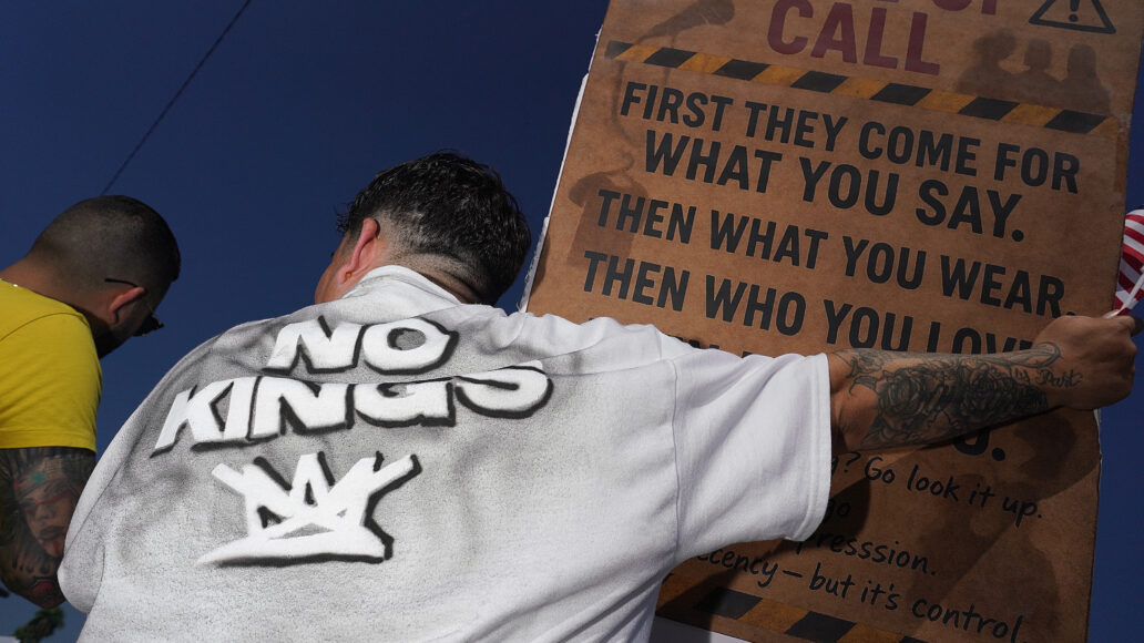 Two men seen from behind; one wears a "No Kings" shirt and holds a brown sign that reads "WAKE UP CALL" and "FIRST THEY COME FOR WHAT YOU SAY. THEN WHAT YOU WEAR. THEN WHO YOU LOVE." He also holds a small American flag.