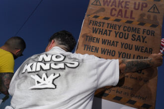 Two men seen from behind; one wears a "No Kings" shirt and holds a brown sign that reads "WAKE UP CALL" and "FIRST THEY COME FOR WHAT YOU SAY. THEN WHAT YOU WEAR. THEN WHO YOU LOVE." He also holds a small American flag.