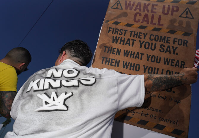 Two men seen from behind; one wears a "No Kings" shirt and holds a brown sign that reads "WAKE UP CALL" and "FIRST THEY COME FOR WHAT YOU SAY. THEN WHAT YOU WEAR. THEN WHO YOU LOVE." He also holds a small American flag.