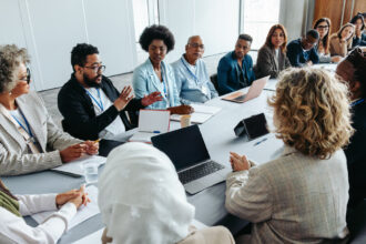 A diverse group of professionals sit around a conference table in a modern meeting room, engaged in discussion during a collaborative meeting, with laptops, notebooks, and coffee cups on the table.